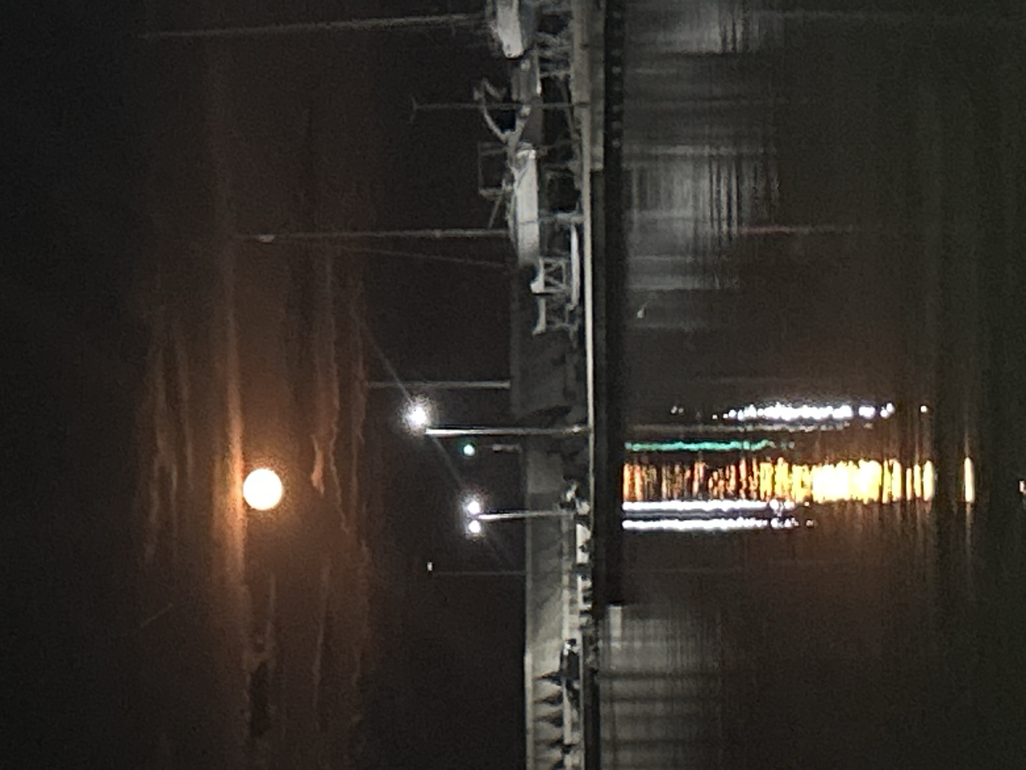 The image depicts a night scene in Burgas, Bulgaria, likely near the sea. A bridge or pier-like structure stretches across the water, marked by repeating lights and a concrete construction style. The moon reflects on the water's surface and illuminates a dark shoreline, potentially indicating the presence of clouds in the night sky. A series of lights, perhaps from the bridge or buildings, appear as a string of luminous dashes reflecting off the water surface.