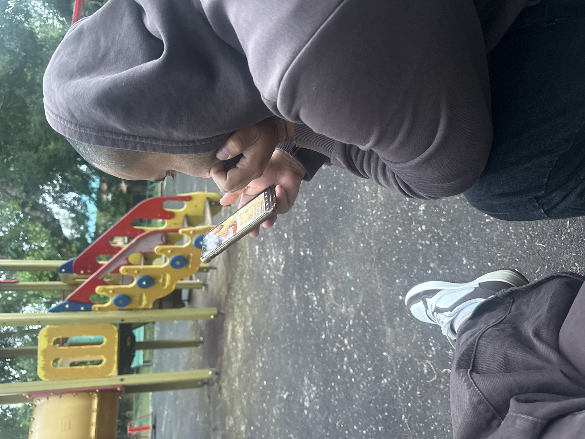 A person, wearing a dark grey hooded sweatshirt and dark pants, sits on the ground, looking at their phone. Their feet, clad in brown and white sneakers, are visible in the lower left corner of the frame. The background reveals a playground with a yellow and red slide structure, and trees with green foliage overhead. The ground is a dark, speckled asphalt. The person appears to be alone. The scene is outdoors, likely in a park or recreational area.