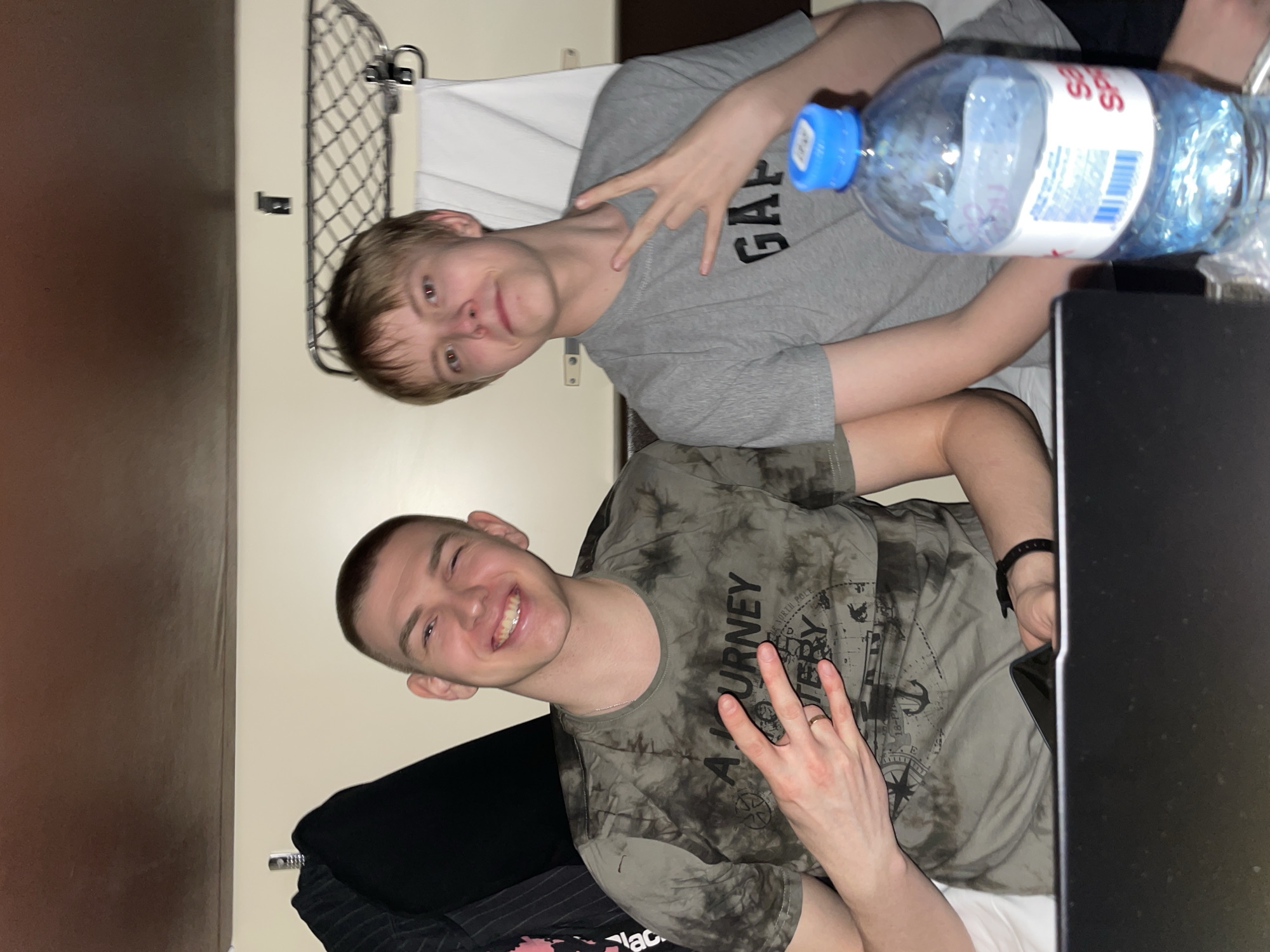 Two young men are posing for a photo inside what appears to be a train car. The man on the left has short brown hair and is wearing a gray t-shirt with "GAP" printed on it. He is holding up two fingers in a peace sign. Next to him is a large plastic water bottle. The man on the right has a shaved head and is wearing a gray and black tie-dye t-shirt with nautical-themed graphics. He is smiling and also holding up two fingers. The background shows a wall with a luggage rack and a black object. Based on the water bottle that says “San Spi”, the location is in the city of Kazan, Russia.