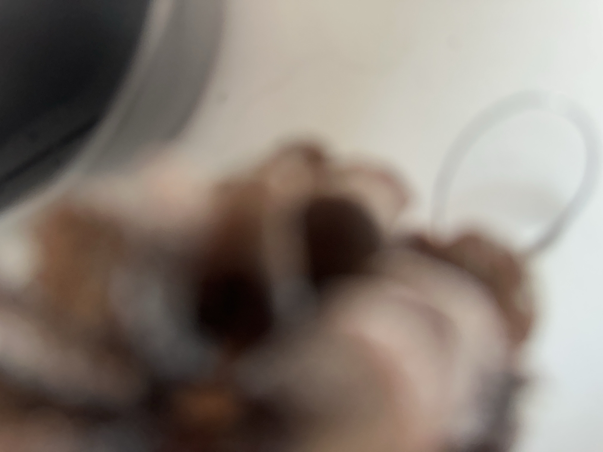 The image is a close-up, out-of-focus shot of a person's head and hand, with a white object in the foreground. The person appears to have dark hair and fair skin. The hand is holding a white, loop-shaped object, possibly a bracelet or a ring. The background is blurred, showing hints of brown and dark gray, suggesting an indoor environment. There are no discernible landmarks or context that would place this image in Belgrade, Serbia.