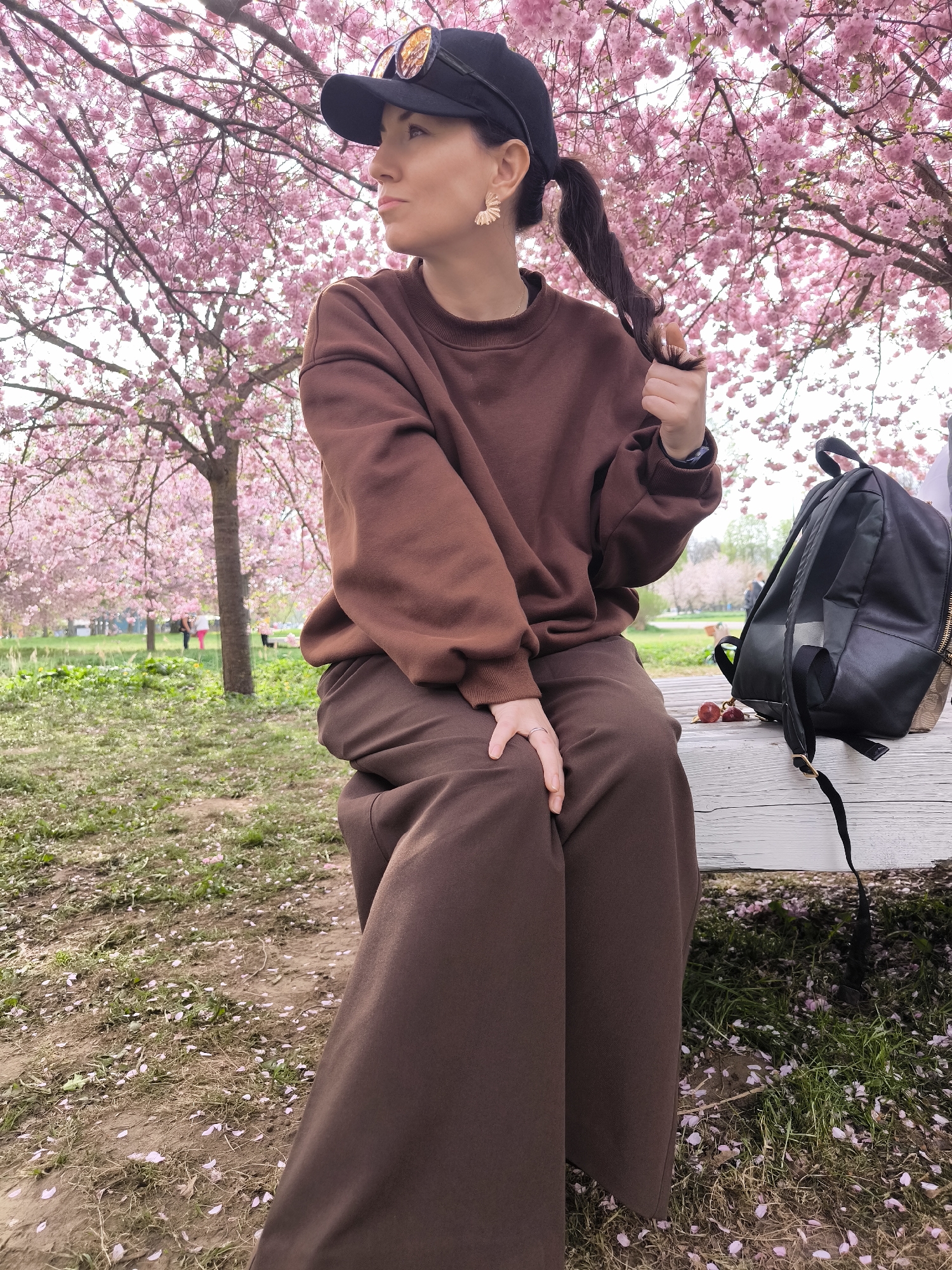 A woman is sitting on a white bench in a park, presumably in Riga, Latvia. She is wearing a brown sweatshirt and matching pants, along with a black baseball cap with sunglasses perched on top. Her dark hair is pulled back in a ponytail. The woman is looking to the left and appears to be holding the end of her ponytail. Next to her on the bench is a black backpack and some cherries. The ground around the bench is covered in fallen pink flower petals from the cherry blossom trees surrounding the area. In the background, more cherry blossom trees and green grass are visible, with a few people walking in the distance.