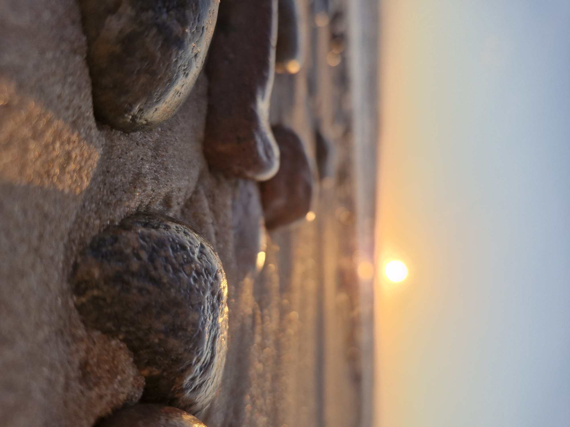 A low-angle, close-up shot captures smooth, wet stones resting on sand at the beach during sunset. The sun is a bright, blurred disc low on the horizon, casting a warm glow over the scene. Waves are visible in the distance, blurred by the shallow depth of field. The sand is damp and reflects the sunlight, creating bokeh effects. The overall impression is one of tranquility and natural beauty, suggestive of a serene moment on the coast of Klaipėda, Lithuania.