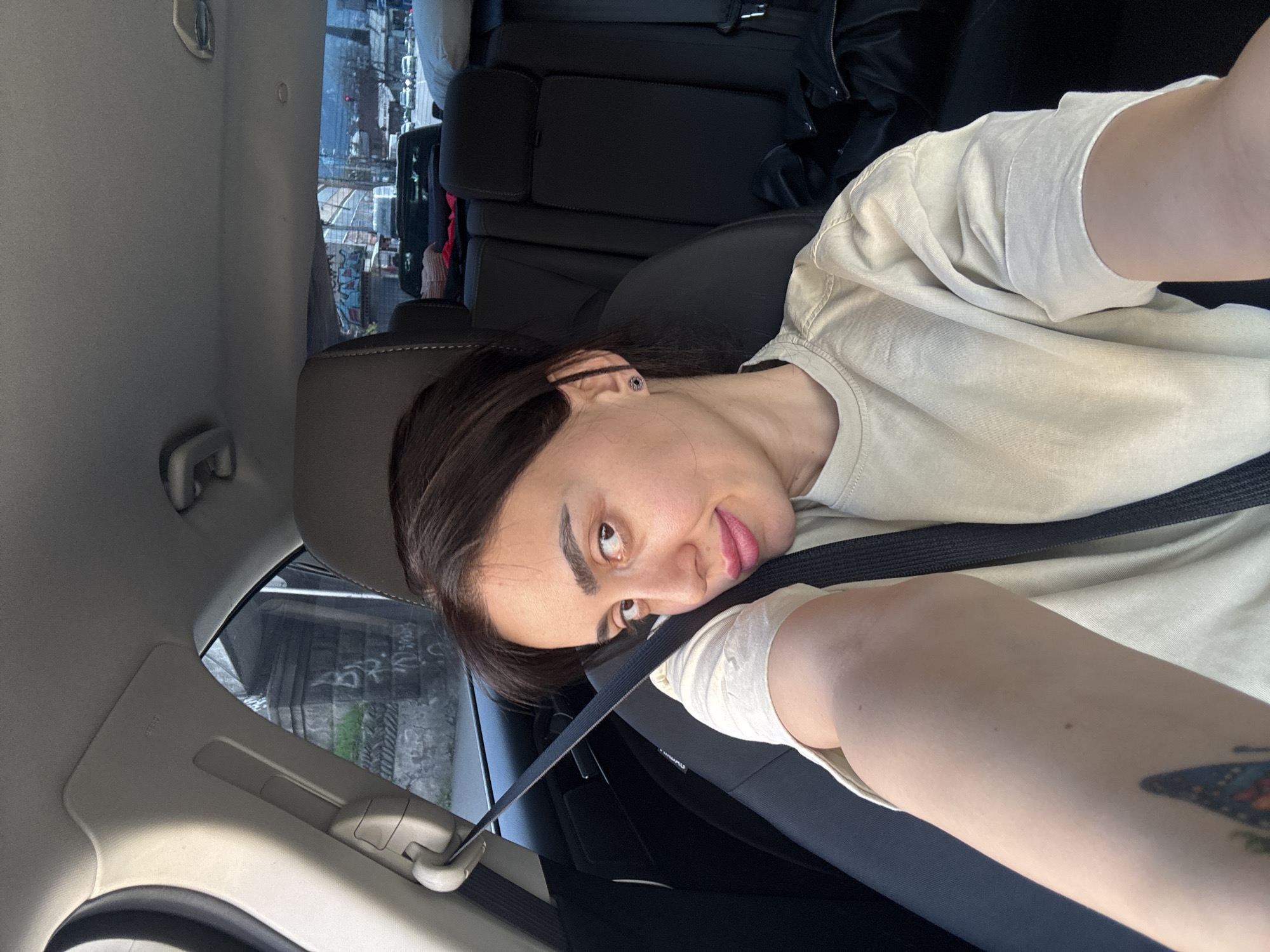 A person with dark hair is seated in the driver's seat of a vehicle, wearing a light-colored t-shirt and a seatbelt. The vehicle is moving on a street with visible graffiti on a stone wall outside the window. The photograph was taken in Tbilisi, Georgia.
