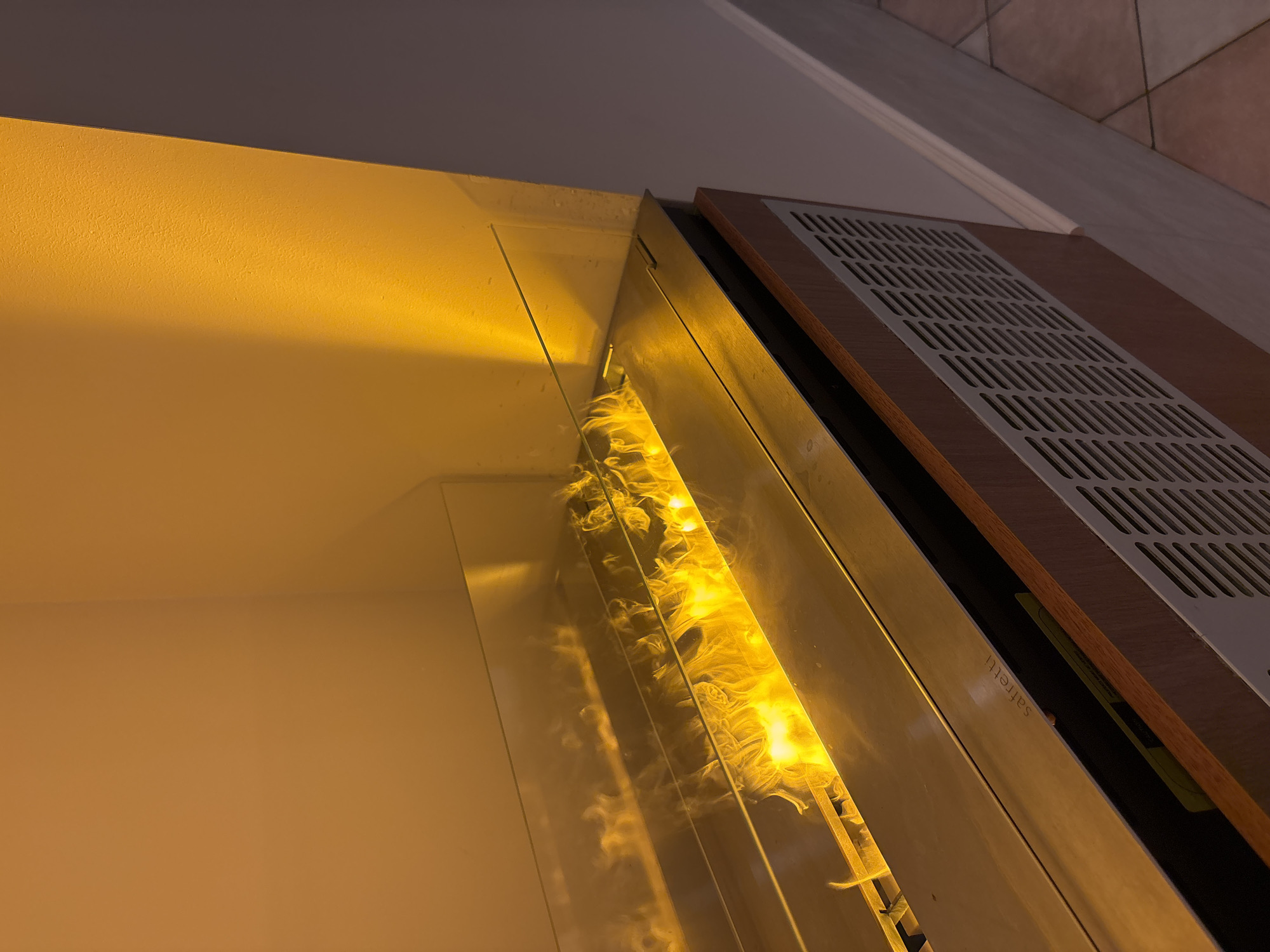 This is a close-up, eye-level, indoor shot of a modern bioethanol fireplace with a light wood veneer surround and a stainless steel grate. The fireplace is lit, producing bright yellow and orange flames and a wispy white smoke effect. The flames are contained by a clear glass barrier. A subtle light wood veneer panel with the word "safretti" is visible on the left side of the fireplace. The background consists of a warm yellow wall and a grey wall. The scene is located in Razliv, Bulgaria.