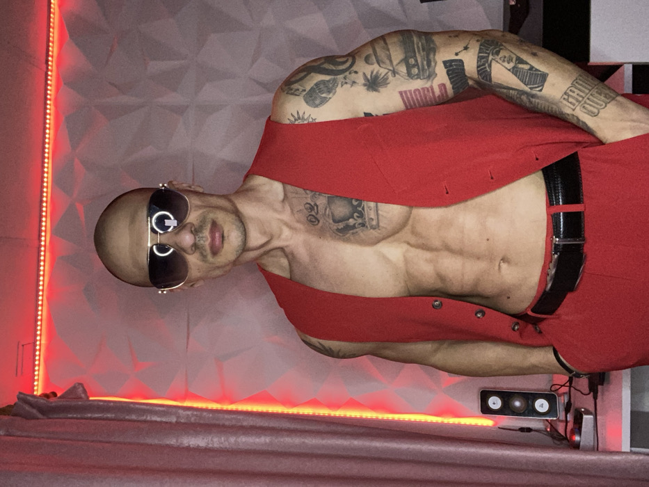 A bald adult male stands facing forward in an indoor setting in Pleven, Bulgaria. He wears aviator-style sunglasses with gold-colored frames and dark lenses, an unbuttoned red sleeveless vest, and red trousers with a black belt. His left hand is in his trouser pocket. The subject's torso is bare, revealing a muscular physique and visible abdominal muscles. Tattoos include a crown design with "02" below it on his left pectoral, and a full sleeve on his left arm featuring designs such as a car, a palm tree, a microphone, an Egyptian profile, and text reading "WORLD DOMINATION" and "GAME OVER." The background consists of a wall covered in white geometric, textured panels, illuminated by red LED strip lighting along the top and left edges. A light-colored curtain is visible on the far left. On a white surface to the left, a vertical electronic device or speaker with two circular elements and associated cables is partially visible. A dark surface is partially visible on the right.