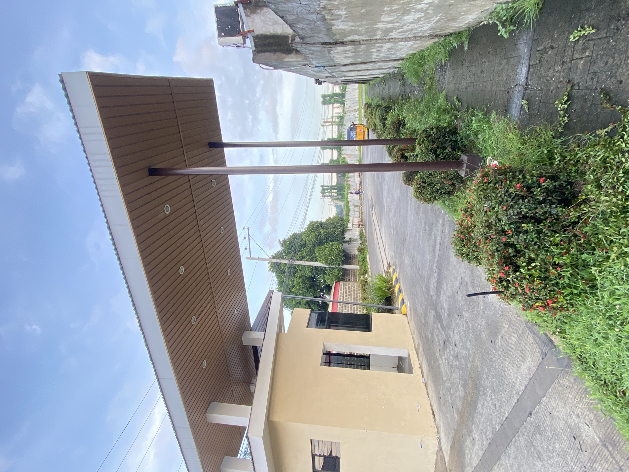 The image depicts an outdoor scene, likely an entrance or gate area, rotated 90 degrees clockwise. In the foreground, a prominent light-colored building, possibly a guardhouse or a small commercial structure, stands on the left. It features dark-framed windows and is partially covered by a broad, light-brown awning with horizontal slatted panels and recessed circular lights on its underside. Two brown support poles for the awning extend downwards from the canopy.
To the right of the building, a paved concrete road or path stretches into the distance, bordered by lush green vegetation. This vegetation includes dense bushes with small red flowers (resembling Ixora) and taller grasses growing alongside the path and a concrete wall. Further along the road, a yellow tricycle, a common mode of transport in the Philippines, is visible. A small, blurry human figure appears to be near the tricycle.
The background reveals a partly cloudy sky, with power lines and utility poles traversing the scene. A concrete wall with what appear to be air conditioning units mounted high up is visible above the vegetation line. Large green trees are also seen in the mid-ground. The ground features areas of cracked concrete and a section with yellow and black stripes, typically indicating a curb or warning area. The overall setting suggests a residential or commercial complex entrance in a tropical environment, consistent with the given location context of Davao City, Philippines. The time of day is daylight, under a bright, though not entirely clear, sky. There is no visible text.