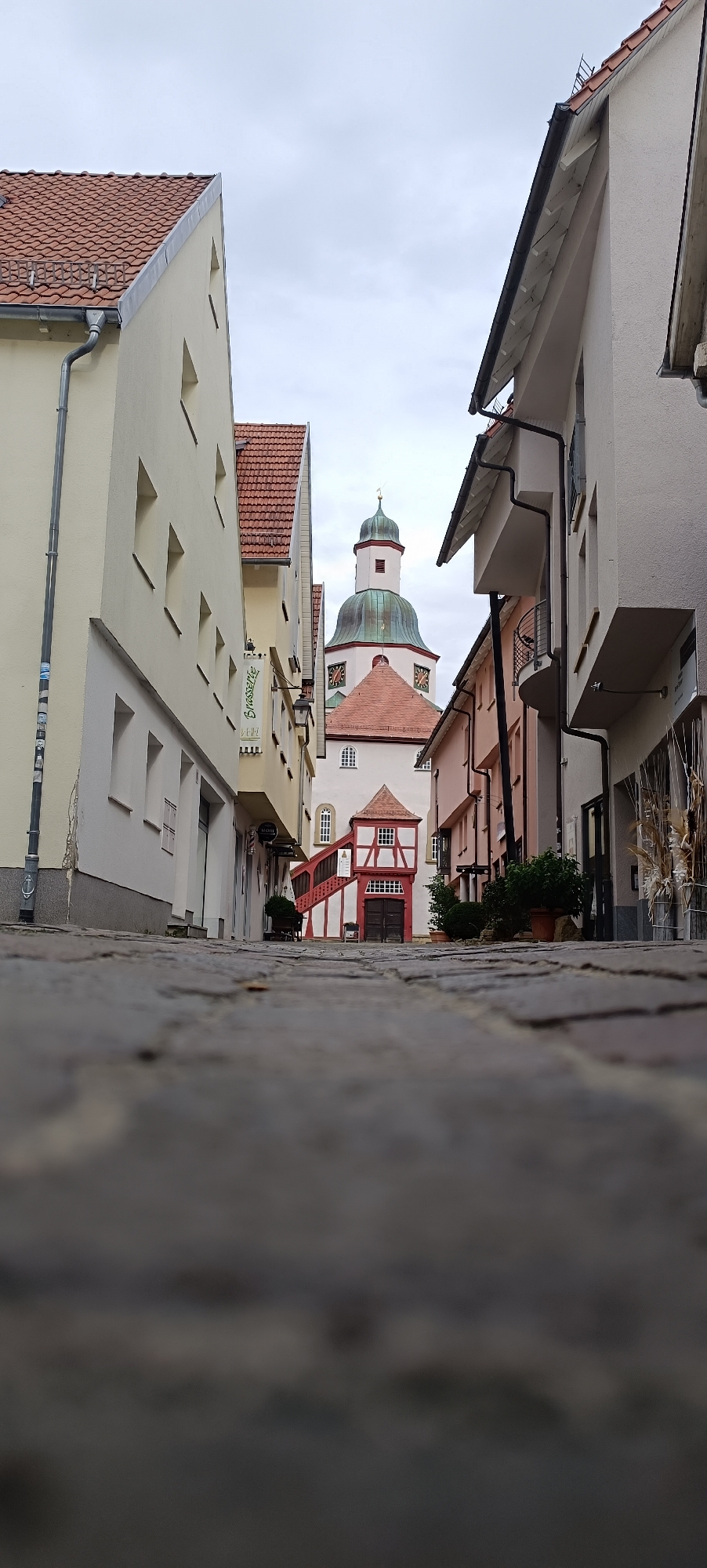 The image depicts a view up a cobblestone alley in Winnenden, Germany, leading to a church with a prominent green dome and clock faces. On the left, a pale yellow building with a red tile roof features several rectangular windows. Further down the alley, a building with a red and white timber-framed facade has an external staircase leading to its entrance. On the right, modern buildings with white and pink facades, featuring balconies and dark gutters, line the alley. The sky is overcast and the overall lighting suggests it is daytime. The image is taken from a low perspective, with the foreground dominated by the textured cobblestones of the alleyway.