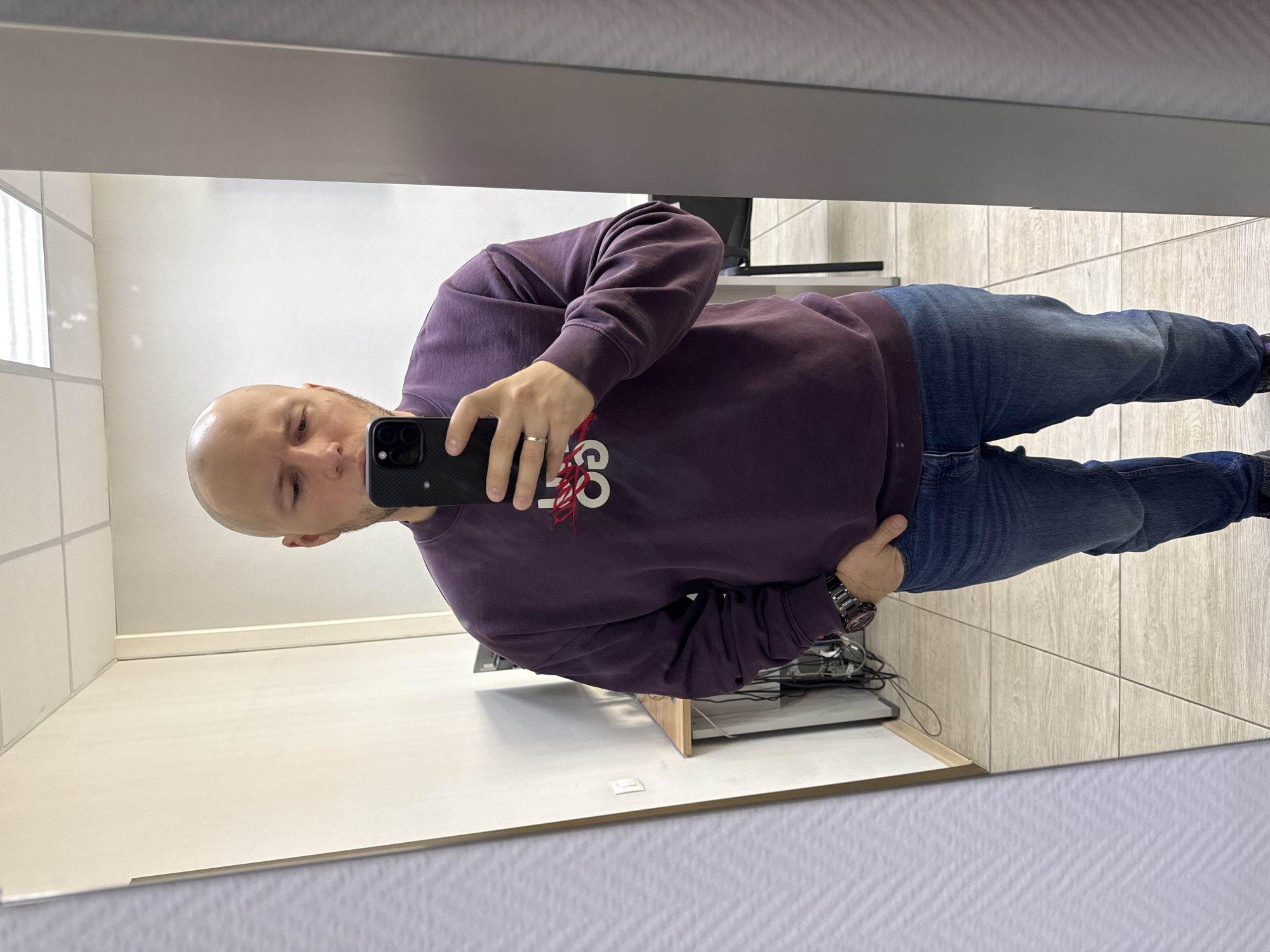 A man stands in front of a mirror, taking a selfie. He is bald, wearing a burgundy sweatshirt and blue jeans. He holds a black smartphone in his right hand. A silver ring is visible on his left hand. The setting appears to be an office or workspace. A desk with electronic equipment and cables is visible behind him. The walls are white, and the floor is tiled with beige-colored tiles. The location is possibly in Krasnoyarsk, Russia.