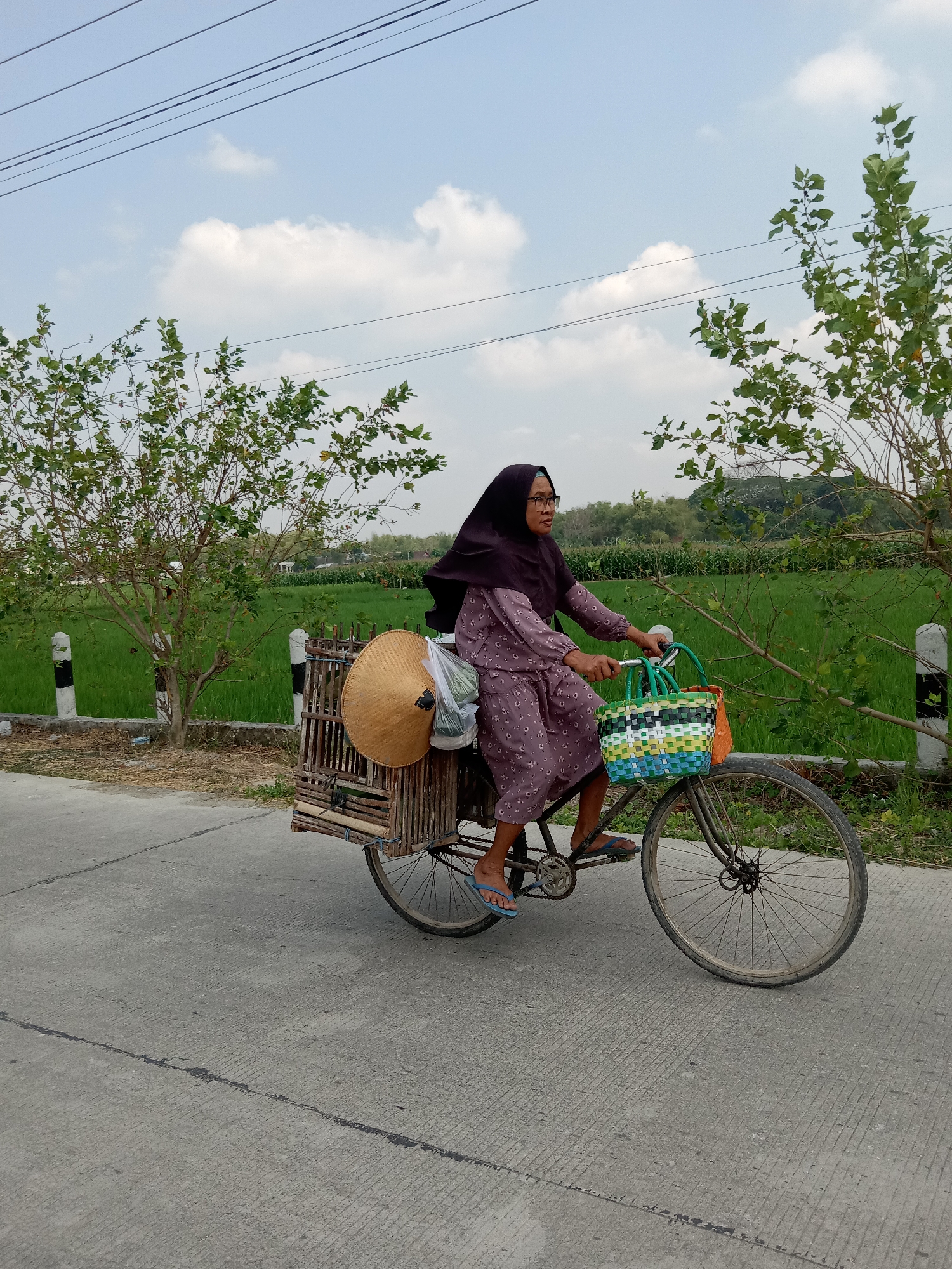 A woman wearing a purple hijab and a patterned dress rides a bicycle on a paved road in Sidobandung, Indonesia. The bicycle has a wooden basket attached to the back carrying a woven hat and plastic-wrapped items. In front of her, a colorful woven basket is attached to the handlebars. The woman is wearing glasses and blue flip-flops. She is looking to her right. The road is bordered by a dry grassy area and a vibrant green field. Several trees with green leaves are visible on either side of the road. A utility pole with black and white markings is also present. The sky is blue with scattered white clouds, and power lines are visible overhead.