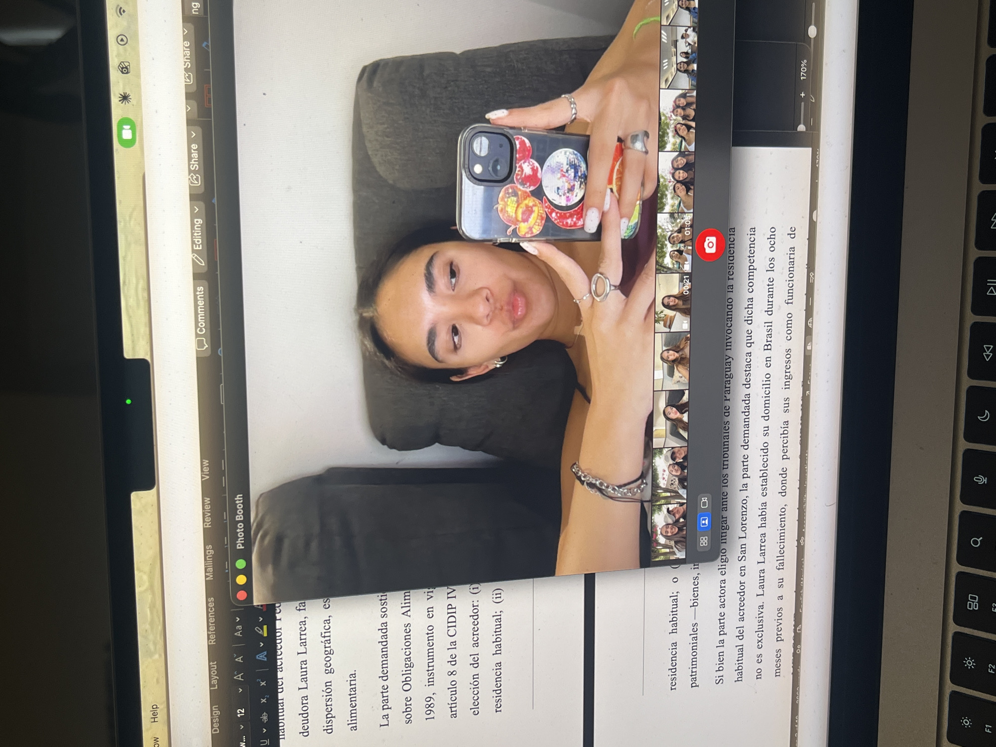 The image shows a laptop screen displaying a Microsoft Word document. On the right side of the screen, there is a partially visible keyboard. The Word document contains Spanish text related to obligations, alimony, and residency of a debtor named Laura Larrea. Superimposed on the Word document is a Photo Booth window, displaying a woman lying on a dark-colored cushion, holding a phone in front of her face. The phone case is decorated with stickers. The Photo Booth window also shows a side bar of multiple photos taken. The environment appears to be indoors. There is no explicit information to confirm the location to be in the city of San Bernardino Tlaxcalancingo, Mexico.