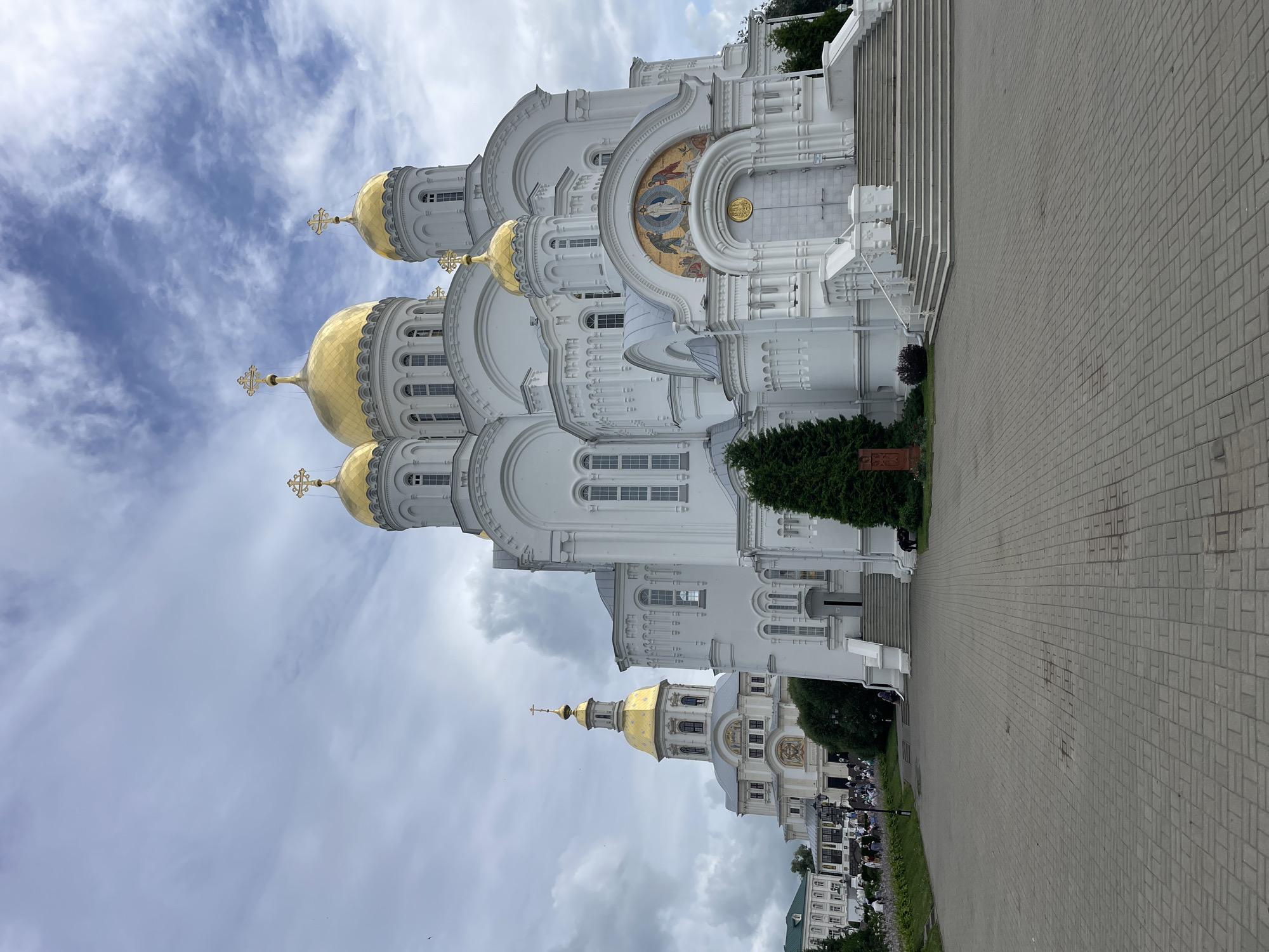 In the city of Diveyevo, Russia, a large, white, multi-domed Orthodox church stands prominently under a cloudy sky. The main structure features numerous golden domes topped with crosses, and decorative golden mosaics above arched entrances. Stairs lead up to the main entrance, flanked by white railings. To the left of the main church, a smaller, taller church with a golden dome and cross is visible, also featuring white architecture and decorative elements. A paved walkway extends in front of the churches, with scattered people in the distance walking on grass and near the buildings. Several trees, including evergreen varieties, are situated around the church grounds. The overall scene depicts a significant religious complex.