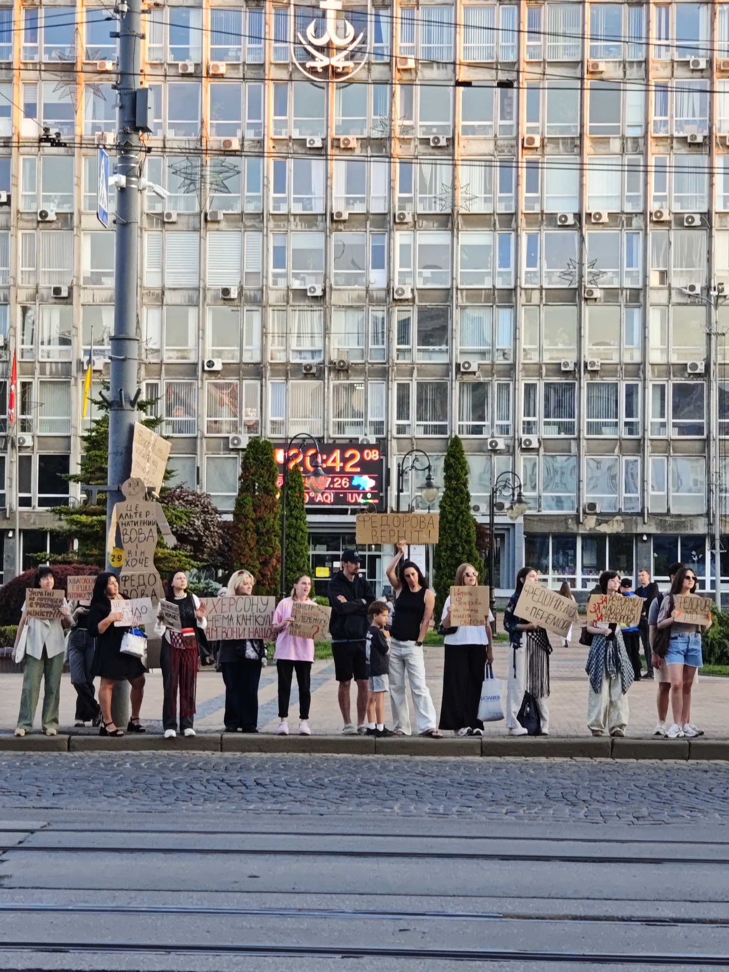 This is a full, eye-level, slightly high-angle shot of a group of people holding protest signs in front of a large, Brutalist-style building in Vinnytsia, Ukraine. The building has a grid of windows and is topped with an emblem. A digital clock displaying "20:42" is visible on the facade. The foreground shows tram tracks on a cobblestone street.
There are at least 13 people visible in the image, appearing to be a mix of men and women, of various ages. Most are standing in a line on a sidewalk, holding signs with handwritten text. Some individuals are looking towards the camera, while others are looking at their signs or the surrounding environment. The people are dressed in casual clothing, with some wearing light-colored tops and pants, and others in darker attire.
The signs are made of cardboard and paper, with handwritten Ukrainian text. Some of the visible text includes "РЕДОРОВА ВЕРНІТЬ!", "ЗА РЕФОРМИ ЗАБОРОНИ НЕ ЗАБОРОНА!", "МІНІСТЕР", and "ХЕРСОНУ НЕМА КАНЦІЯ АМ ВОНИ НАШІ". The signs suggest a protest, likely related to political or social issues in Ukraine.
The environment appears to be an urban setting during the daytime, possibly late afternoon or early evening, given the clock reading and lighting. There are trees and streetlights visible. The overall atmosphere suggests a public demonstration.