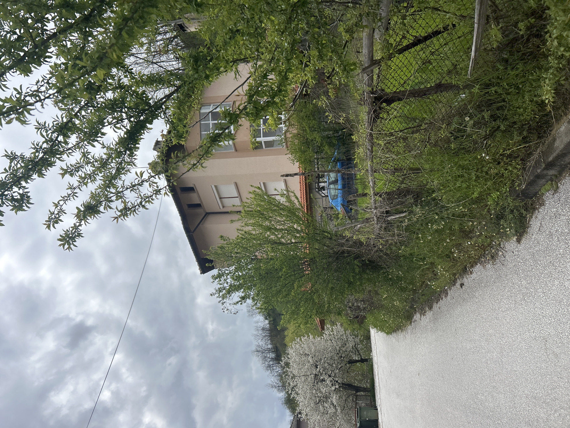 A residential area in Zlatograd, Bulgaria, features a two-story, light-beige building with a red-tiled roof visible through the surrounding trees. The building has white-framed windows, including two windows on the upper level and two smaller windows on the lower level. A chain-link fence borders the property, partially obscured by dense green foliage. A paved road runs alongside the property, and beyond the fence, there's a hill with trees and flowering white trees. The sky is overcast with heavy clouds. The angle of the photo suggests it was taken from a low vantage point, looking upward.