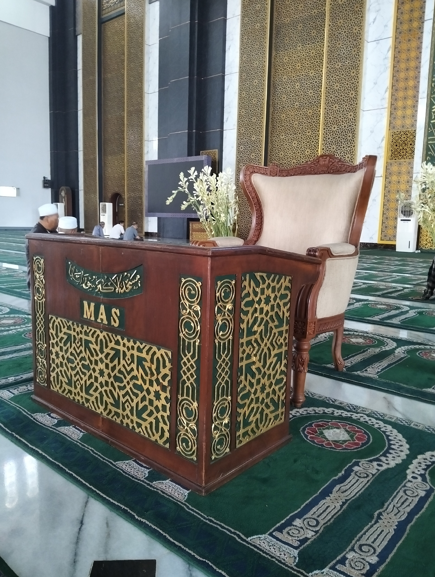 A mosque interior in Surabaya, Indonesia, features a dark brown, ornate wooden podium with gold decorative inlays and the word "MAS" in gold letters. A beige upholstered armchair with dark wood trim sits to the right of the podium. In the background, men in white skullcaps are seated on a green carpet patterned with a red and white floral design. The walls are adorned with vertical gold and black paneling. The carpet on the floor is predominantly green with intricate patterns.