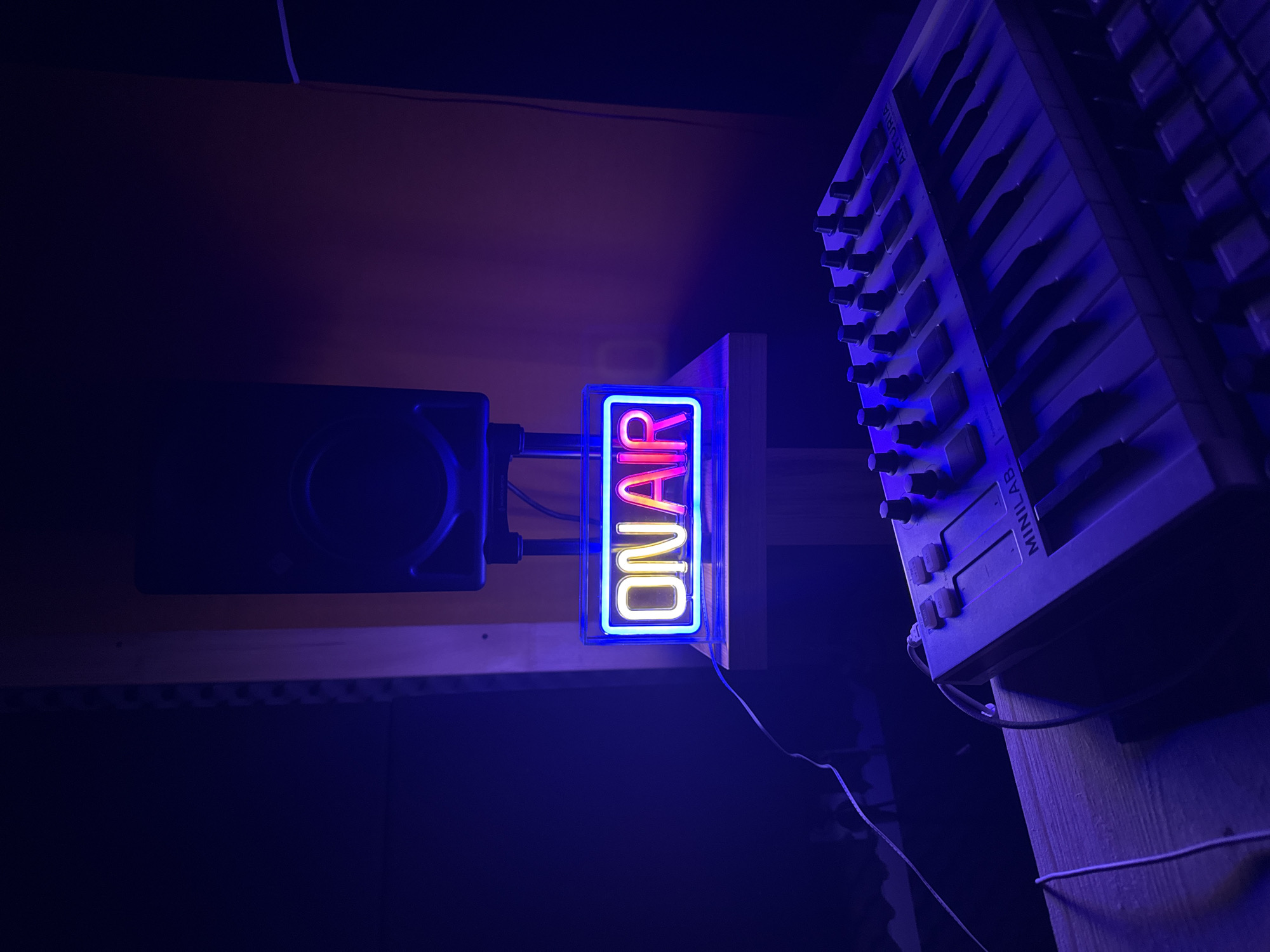A low-angle, medium shot indoors shows a blue and red "ON AIR" sign, a black speaker, and a keyboard, possibly a MIDI keyboard, attached to a wooden surface. The environment appears dark with blue lighting. The location is unspecified and has no apparent connection to Plovdiv, Bulgaria. The image focuses on the studio equipment.