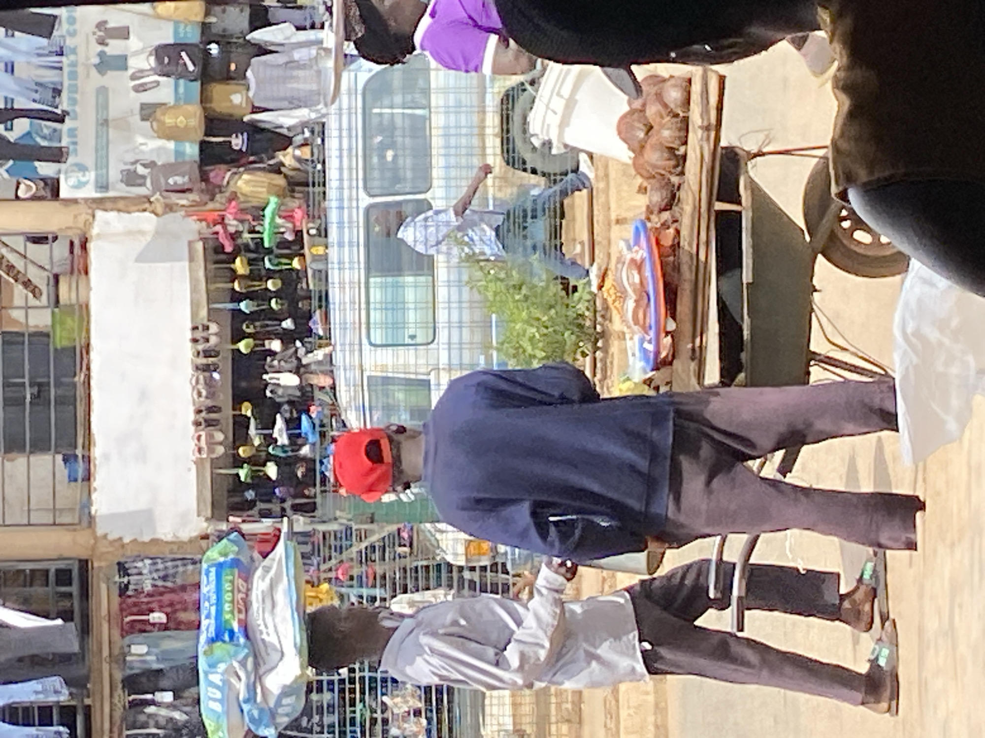 A street market scene in Bukuru, Nigeria, is depicted in this outdoor, eye-level shot. The focus is on two men in the foreground. The man on the left, wearing a light gray collared shirt and dark gray pants, has a stack of items wrapped in plastic bags balanced on his head and is looking to his right. He is wearing sandals. The man on the right is wearing a dark blue sweatshirt and dark gray pants, with a red baseball cap on his head, facing away from the camera and towards the man on the left.
Behind the men, a bustling market is visible with stalls displaying various goods. Clothing items hang from racks, and footwear is displayed. A light blue and white checkered van is parked in the mid-ground, with a person in a light blue plaid shirt walking in front of it. A woman in a purple shirt with white trim is partially visible on the right edge of the image.
In the immediate foreground, a wooden cart with a wheel holds food items. On the cart, there are plates of food and a pile of brown, round objects, possibly fruits or nuts. A white bucket is also present near the cart. The ground appears to be a dirt or sandy surface. The lighting suggests it is daytime, with shadows indicating a sunny environment. The overall impression is of a busy commercial area.
