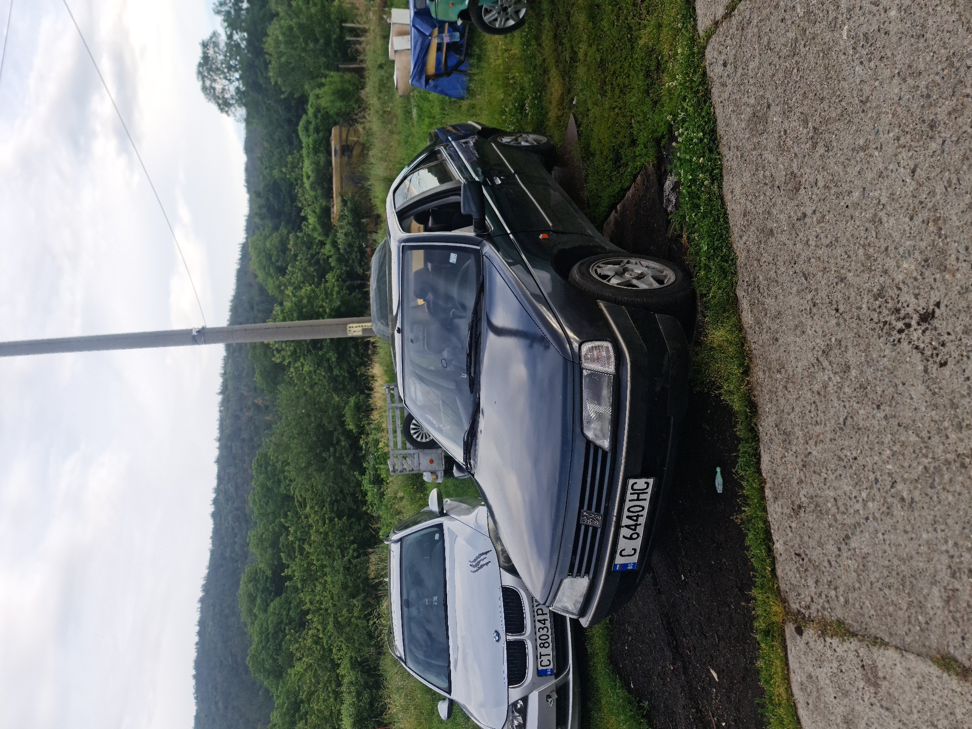 This outdoor image, taken in Buzovgrad, Bulgaria, captures a semi-rural scene featuring two parked cars, a power pole, and a hilly, tree-covered landscape under a partly cloudy sky.
In the foreground, a dark blue or black Peugeot sedan (likely a 406 model) is parked on a grassy area, facing towards the right side of the frame. Its front end, including the headlights, grille with the Peugeot emblem, and bumper, is clearly visible. The car displays a Bulgarian license plate "C 6440 HC," which includes the Bulgarian flag and "BG" country code.
Behind the Peugeot, a silver BMW wagon or SUV is partially visible, also facing right. Its front grille with the BMW emblem, headlights, and part of the hood are seen. This vehicle also bears a Bulgarian license plate, partially legible as "CT 8034 PY," along with the Bulgarian flag and "BG" identifier.
To the right of the parked cars, a concrete path or driveway extends into the distance. The ground immediately surrounding the vehicles is covered in green grass, with some patches of darker soil or mud. A light blue plastic bottle is discernible on the grass near the Peugeot's front bumper.
On the far left, a utility pole stands tall, with a small, illegible yellow-and-white sign attached to it. In the background, beyond the vehicles, dense green trees cover rolling hills, extending across the horizon. Further back in the grassy area, a smaller vehicle or trailer, partially covered with blue tarps, is visible.
The scene appears to be set during the daytime, under natural, somewhat overcast lighting conditions. No people or specific activities are visible, presenting a static depiction of parked vehicles in a natural setting.