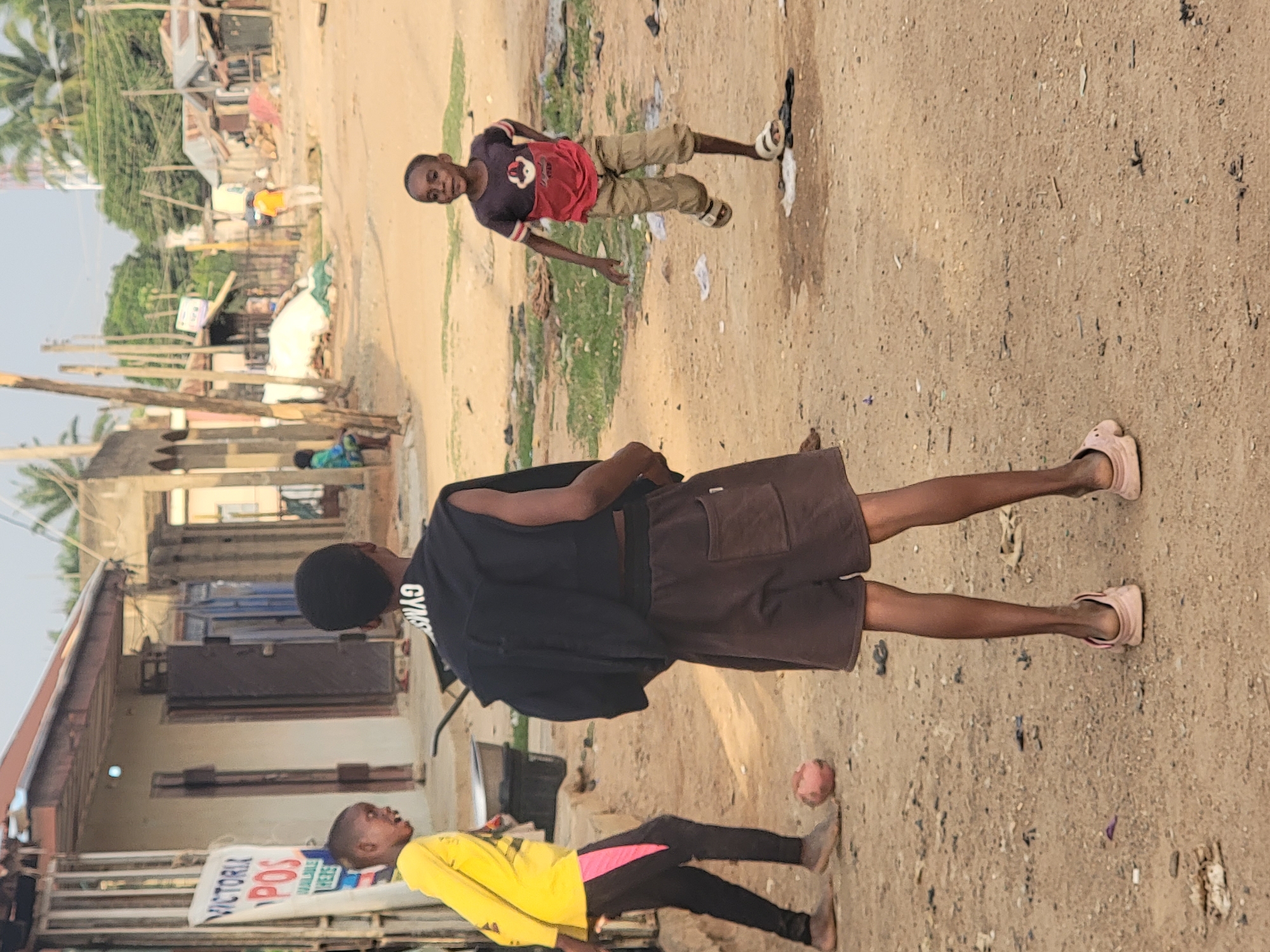 The image captures a street scene in Sabon Tasha, Nigeria, featuring three children. One child, wearing a yellow shirt with pink and black accents and black pants, stands near a "Victoria POS" sign. Another child, dressed in a burgundy shirt and tan shorts, is positioned further up the street. The third child, in the middle ground, is wearing a black shirt, brown shorts, and pink sandals. The street appears to be a dirt road with some greenery on the side. Buildings and structures are visible in the background.