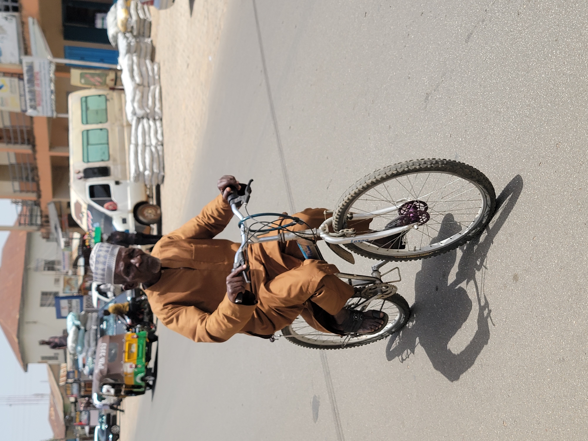 In Jos, Nigeria, an elderly man wearing a tan shirt and a white patterned cap is riding a bicycle on a paved road. Buildings and parked vehicles, including a white van loaded with sacks, are visible in the background. The scene appears to be a busy street within the city.