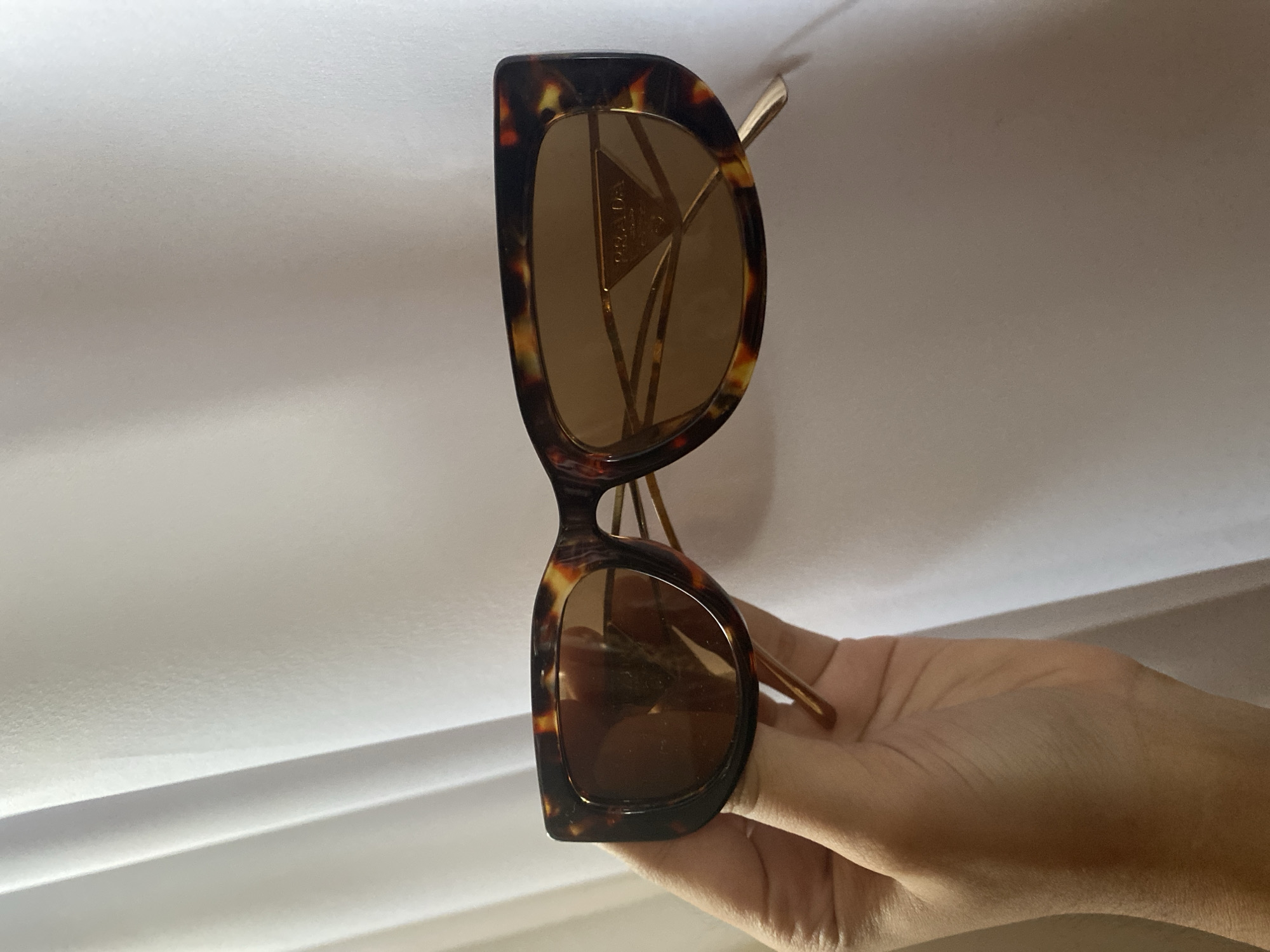 A pair of Prada sunglasses with a tortoiseshell frame and brown lenses is held in a hand against a background of white fabric. The location is not discernible from the image.