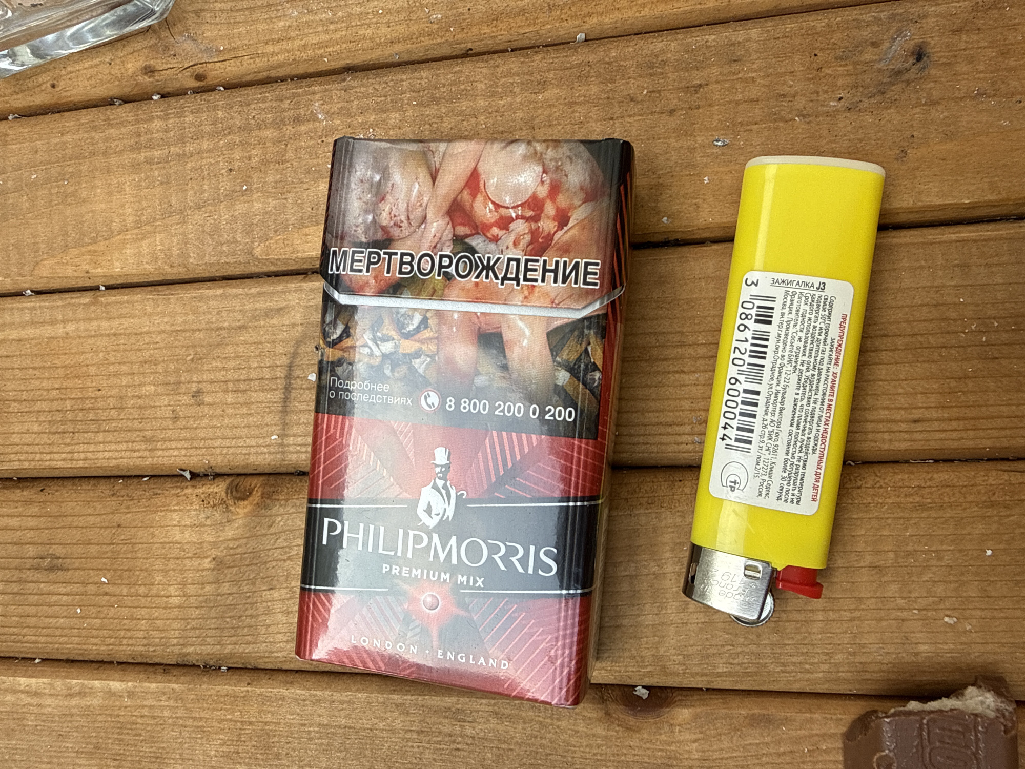 An overhead shot shows a yellow lighter lying on a wooden surface next to a pack of Philip Morris cigarettes. The cigarette pack is red and black with an image of a bloody fetus on the right side. The lighter has the text "Zazhigalka" on it, meaning "lighter" in Russian. The wooden surface appears to be an outdoor table. The image is taken from a high angle.