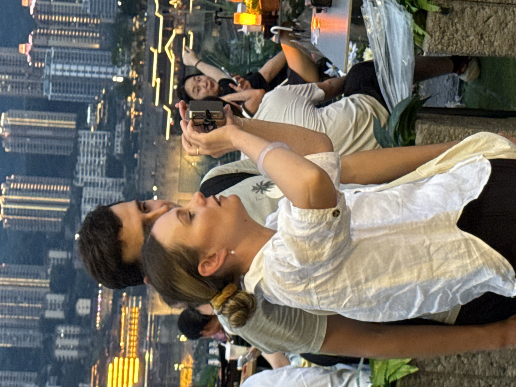 In a nighttime urban setting, likely in Jiangbeicheng Street, China, a woman in a white shirt is in the foreground, holding a cell phone and taking a photo. Her hair is pulled back in a bun, and she is wearing a pearl earring. She is standing next to a man. Behind them, other people are visible, and in the background, illuminated skyscrapers and buildings suggest a bustling cityscape. A table with a lantern is also in the scene.