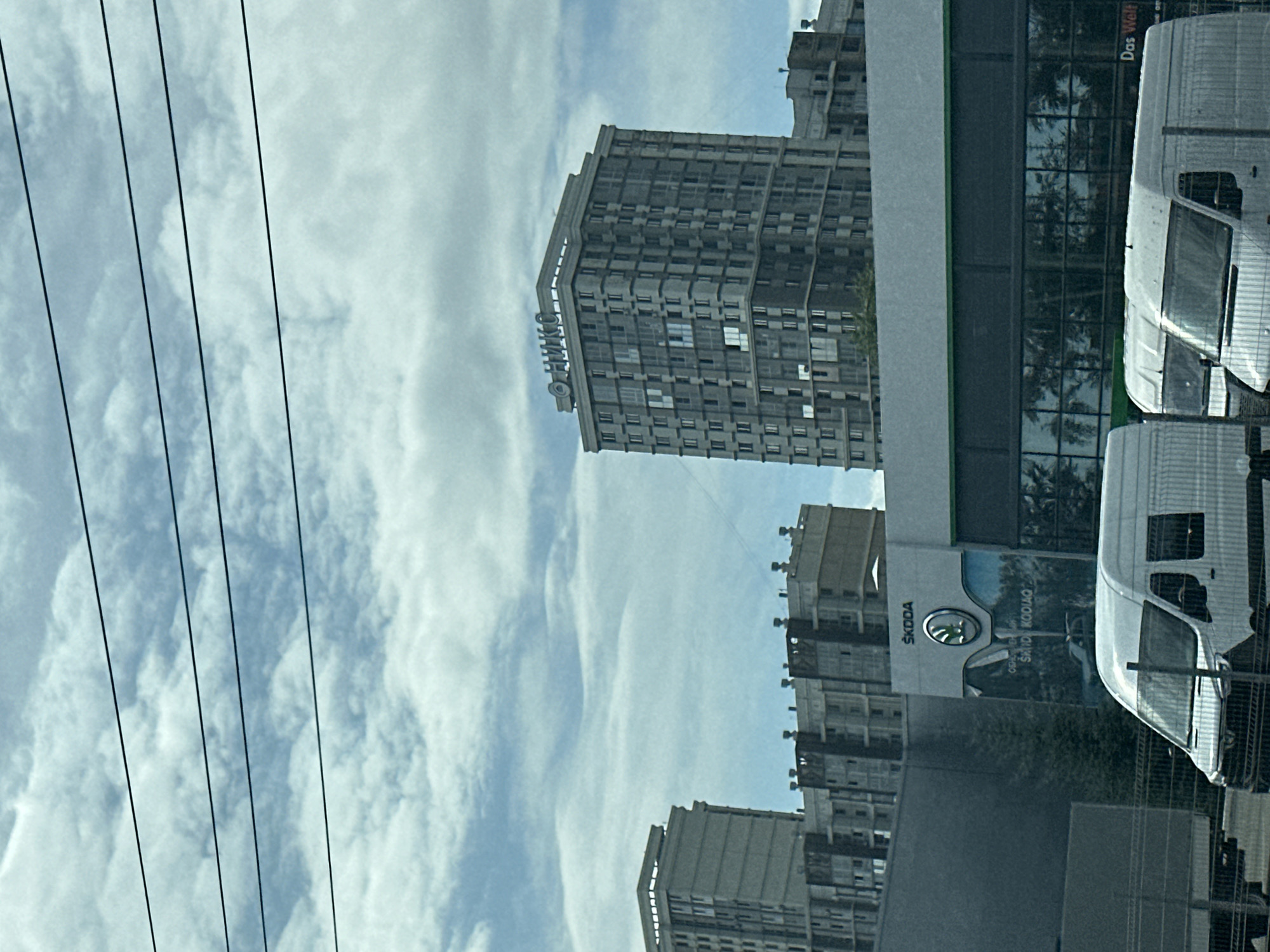 In this street-level, medium-shot photograph taken from a slightly low angle, several buildings and vehicles are visible under a cloudy sky. In the background, two tall, modern apartment buildings dominate the scene. The building on the right is darker gray with many windows and a sign at the top that reads "ОНИКС" in white lettering. The building on the left is lighter gray and appears to be under construction or in a similar style. In the midground, a low-rise building with a glass facade and a green trim runs horizontally across the frame. Above this building, a sign for "SKODA" is visible, with the car brand's logo. The glass facade reflects trees and the sky. In the foreground, two white vans are parked. The van on the left is partially obscured by the frame. The van on the right is fully visible, with its front end facing the camera. The image is captured on a slightly overcast day, with thin, dark power lines stretching across the top of the frame. The setting appears to be an urban environment.