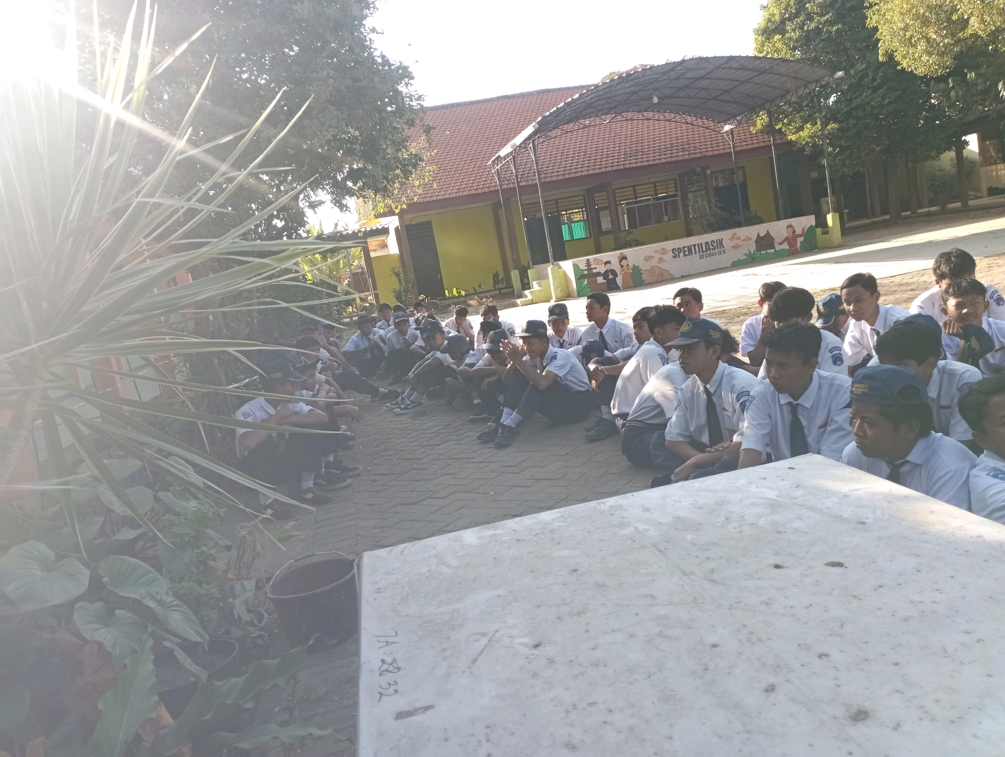 In the city of Banyuurip, Indonesia, a group of students, likely in uniform with white shirts and dark ties, are seated on a paved area in front of a school building. The building features a red tiled roof and yellow walls, with an open-air covered structure in front. Some students are looking towards the right of the frame, while others are looking at something out of frame. A large concrete slab is in the foreground. To the left, a lush green plant with long, spiky leaves is illuminated by bright sunlight, casting lens flares across the image. The environment appears to be outdoors, with trees and foliage visible in the background, suggesting a schoolyard or outdoor gathering space.