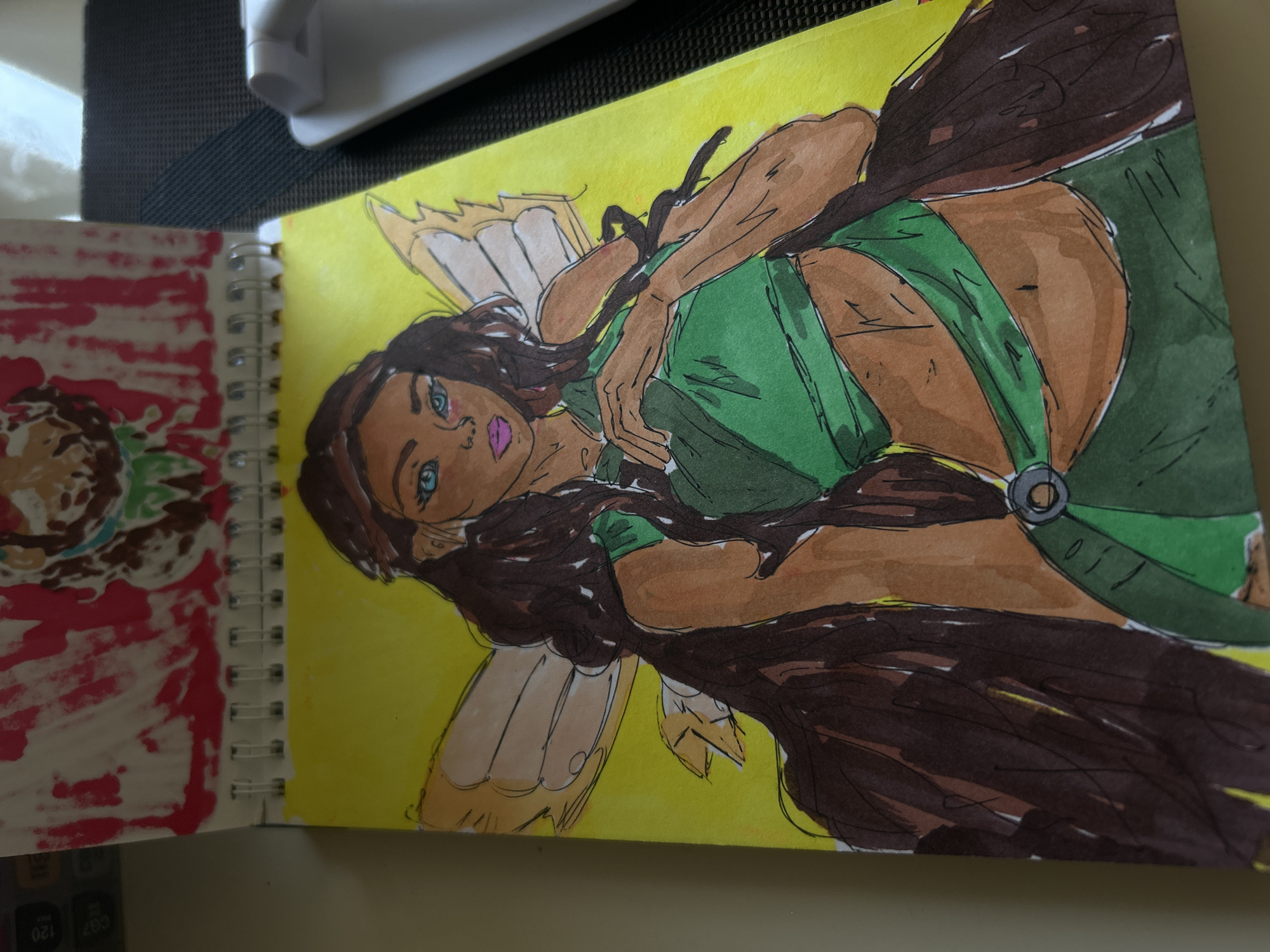 The image displays a drawing of a fairy on a yellow background, situated within a sketchbook. The fairy has long, dark, wavy hair, blue eyes, and is wearing a green outfit. She has delicate wings and her skin tone is medium brown. The drawing is colored with markers, exhibiting visible lines and some slight bleeding of the ink. The sketchbook itself is spiral-bound, with another drawing, primarily in red and pink, visible on the page above. The overall environment suggests an indoor setting, possibly a desk or table, with the corner of a white object, possibly an electronic device, visible in the upper right portion of the frame. There is no indication or context within the image that places it in Khyrdalan, Azerbaijan or any specific city.