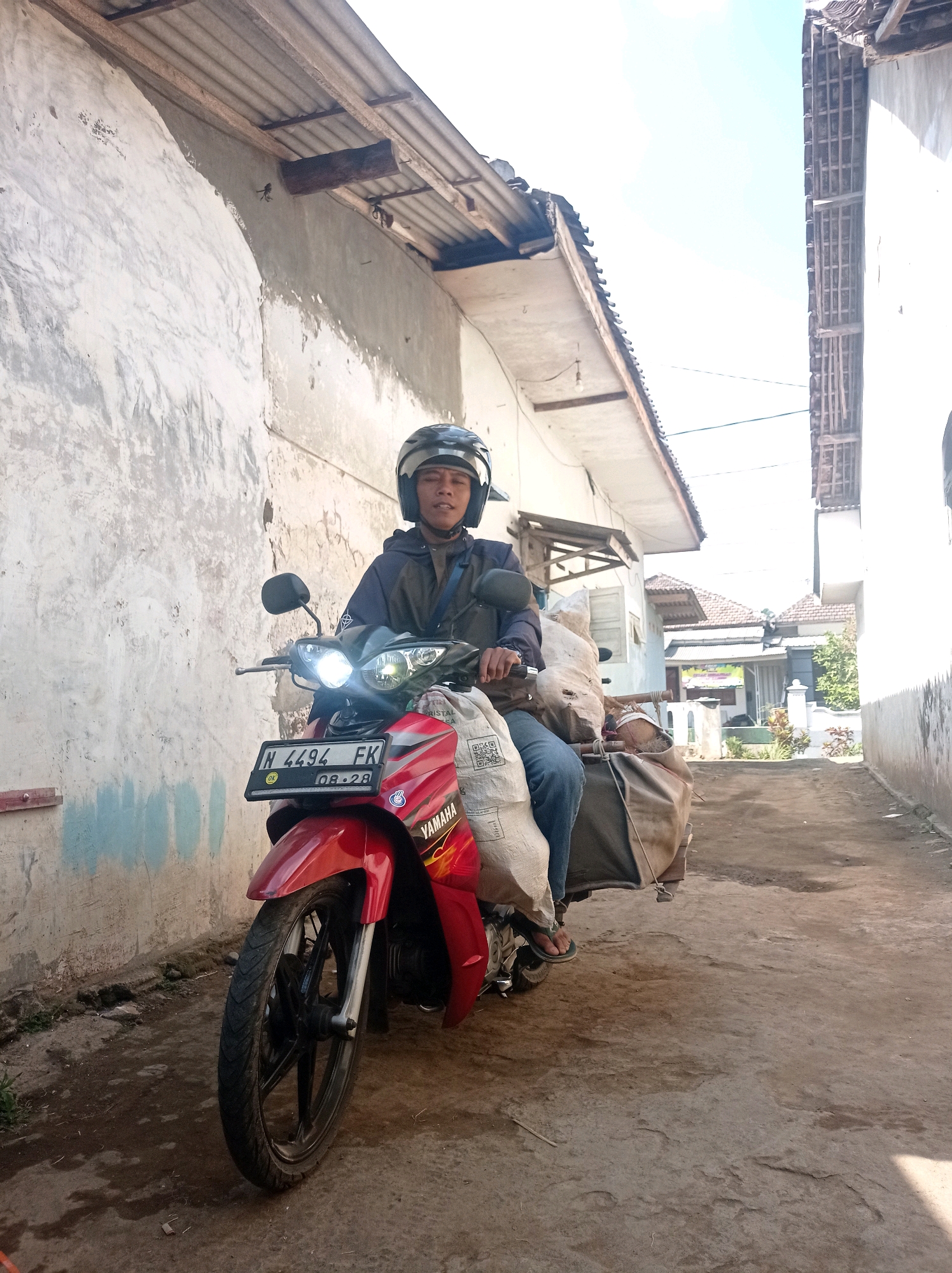 In Purwosekar, Indonesia, a person wearing a black helmet rides a red Yamaha motorcycle. The motorcycle is carrying several large, light-colored sacks strapped to the back. The rider is wearing a dark jacket and blue jeans, with flip-flops on their feet. They are positioned on a dirt path, with buildings and a wall lining either side. The wall on the left is weathered and textured, with some blue paint visible. The buildings on the right appear to be houses with tiled roofs. The sky is bright and clear. The license plate on the motorcycle reads "N 4494 FK 08.28".