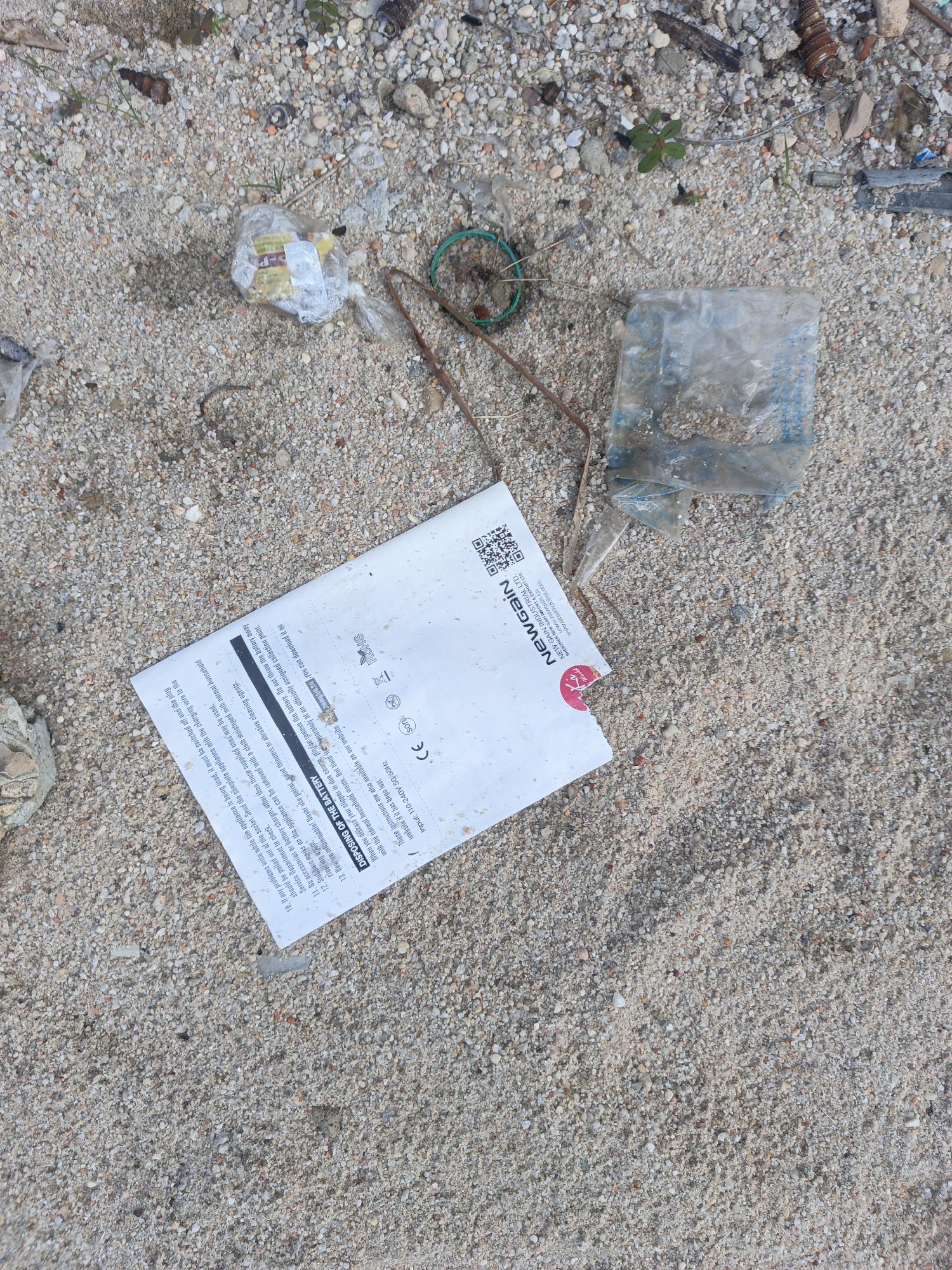 The image shows a sandy, pebbly ground littered with trash. Prominently featured is a partially torn paper sheet with the brand "NEWGAIN" printed on it, accompanied by recycling and CE symbols. The sheet also contains instructions for disposing of a battery. Near the paper, there are plastic bags, one clear and another containing what appears to be food packaging. A rusted metal piece and a green plastic ring are also scattered around. The environment appears to be outdoors, possibly a beach or an unkempt urban area. There are no people visible in the frame.