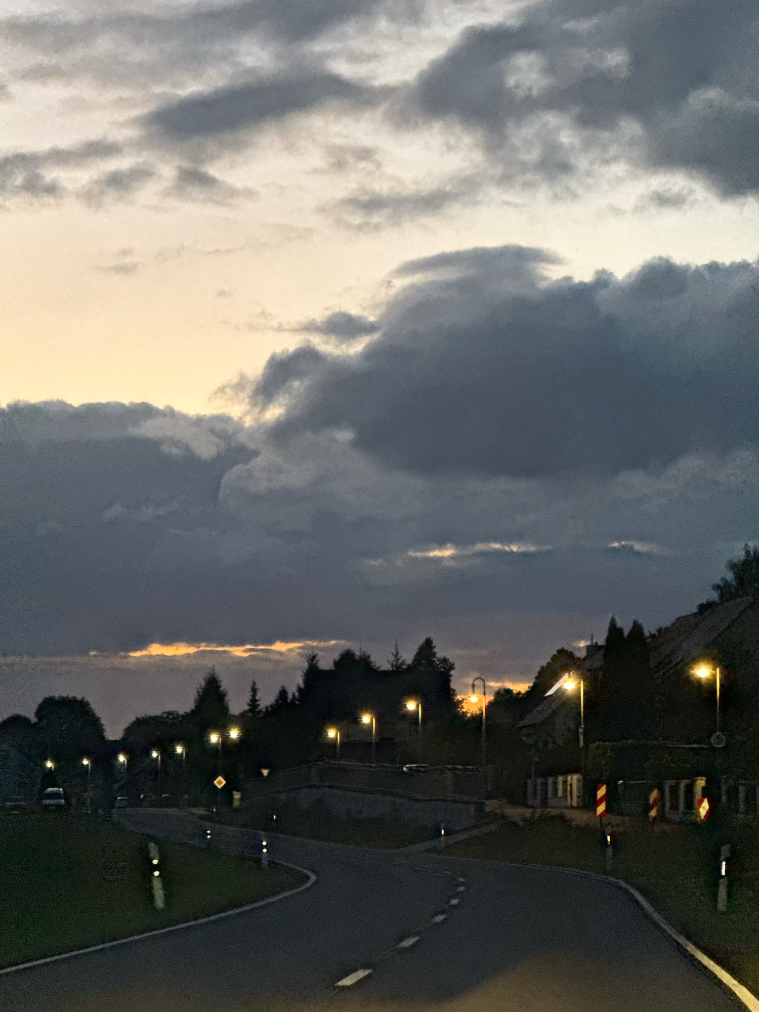 The image shows a rural road in Werther, Germany, at dusk. The sky is filled with dark, dramatic clouds, with a sliver of orange light visible near the horizon. The road curves through a landscape of trees and houses. Streetlights are illuminated along the roadside, casting pools of yellow light. Several cars are parked on the left side of the road. Traffic signs and bollards are visible along the road, indicating a curve ahead. The scene conveys a sense of twilight tranquility, with the artificial lights of the town contrasting with the natural elements of the sky and trees.