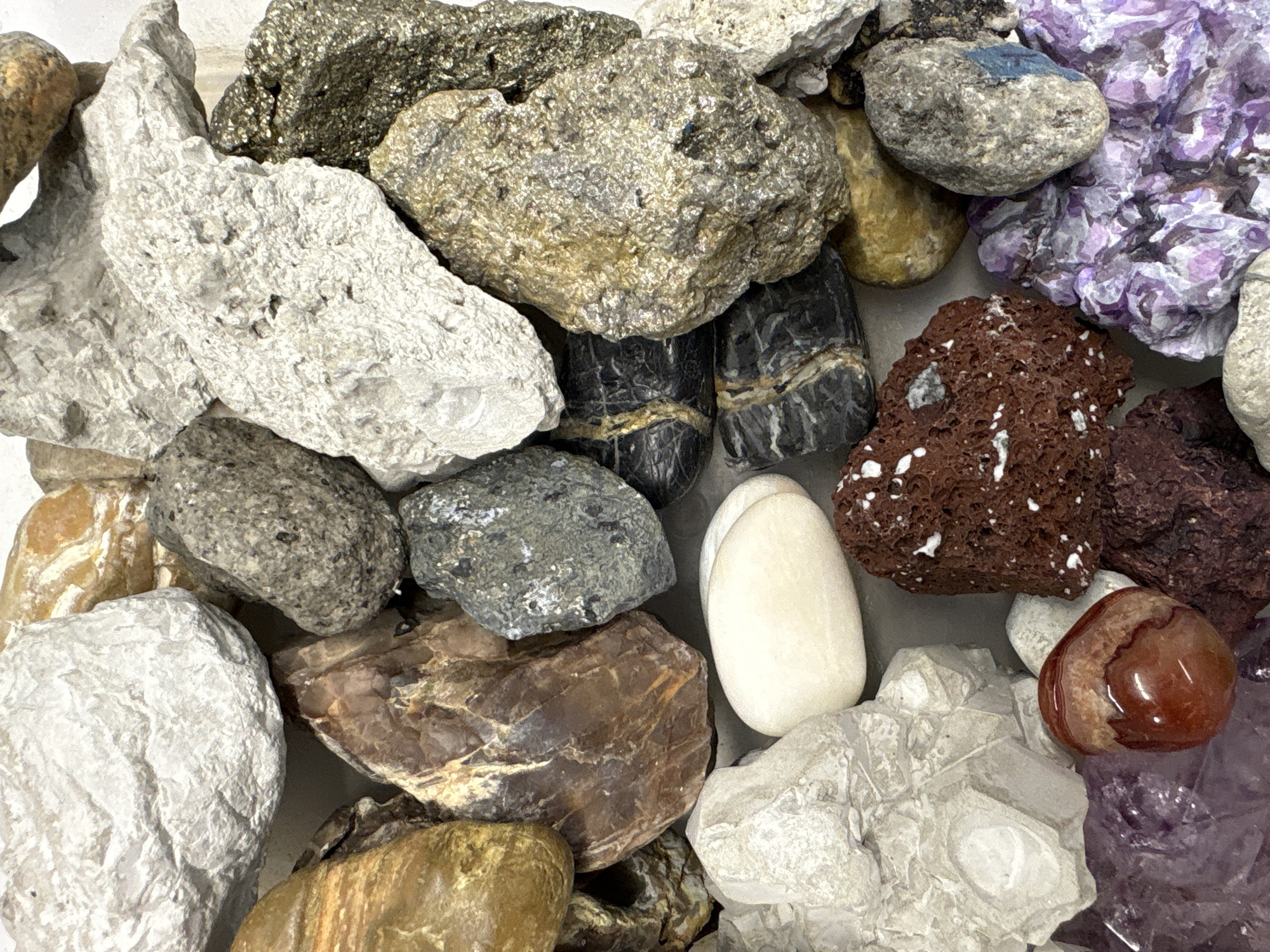 The image displays a collection of various rocks and minerals. The arrangement appears random, with the items overlapping and clustered together. Colors range from grays, browns, whites, and purples. Some rocks show distinct textures such as porous surfaces, veining, and crystalline structures. One rock is a smooth white pebble, while another is a shiny reddish-brown agate. The presence of amethyst suggests the possible locality or source might be related to mineral collecting or a geological display. Location information is unavailable.