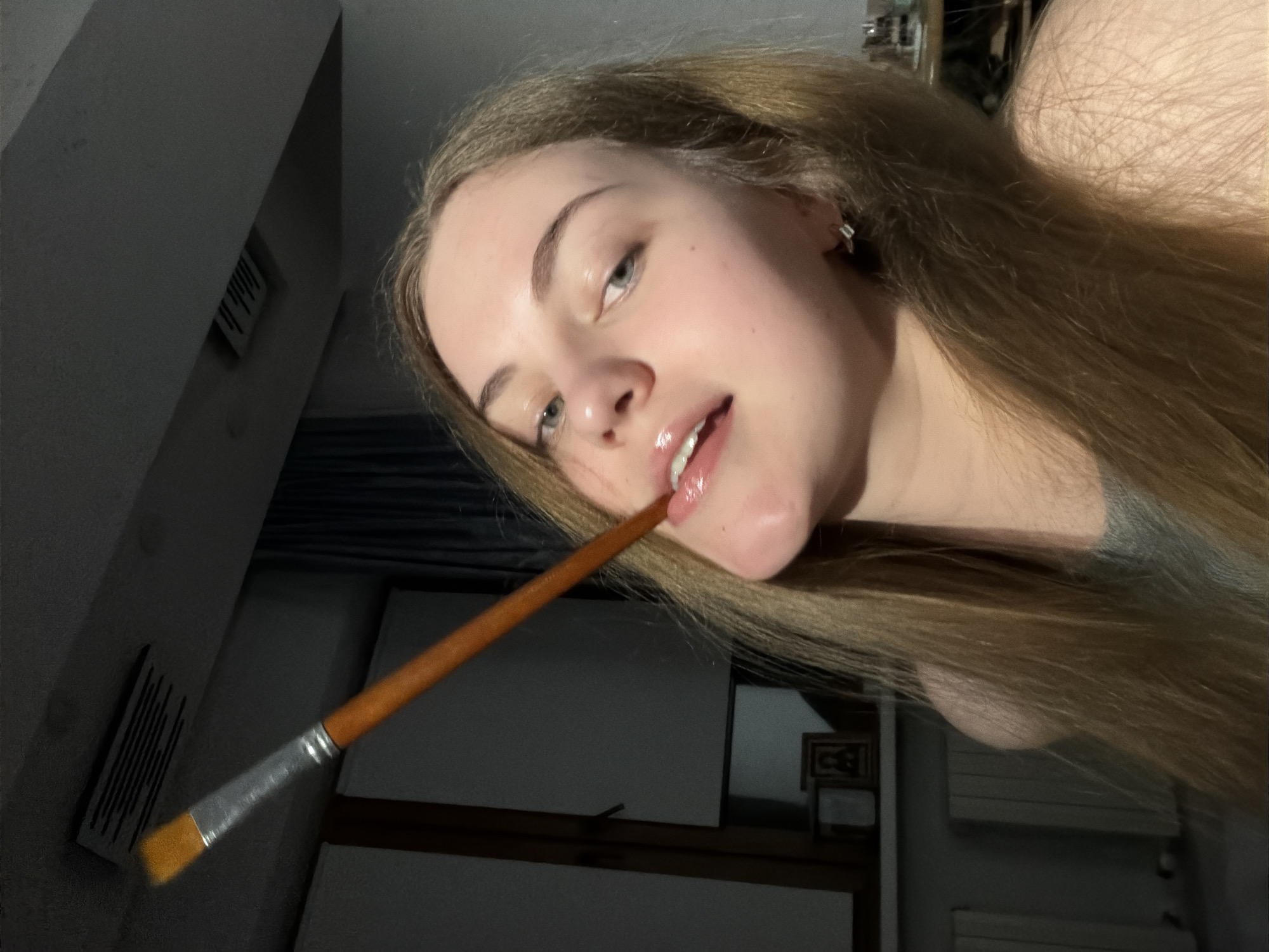 A young woman with long blonde hair is captured in a close-up, low-angle shot. She is holding a paintbrush, the bristles of which are positioned near her lips, and she appears to be biting the handle. Her lips are glossy, and her eyes are open and looking towards the camera. She is wearing a gray shirt and a small silver earring in her right ear. The background is dimly lit and appears to be an indoor setting, possibly a room in Dnipro, Ukraine, with a window on the left and what looks like a radiator below it. A ventilation grate is visible on the ceiling above the woman's head. The lighting suggests the scene is taking place at night or in a dimly lit room.