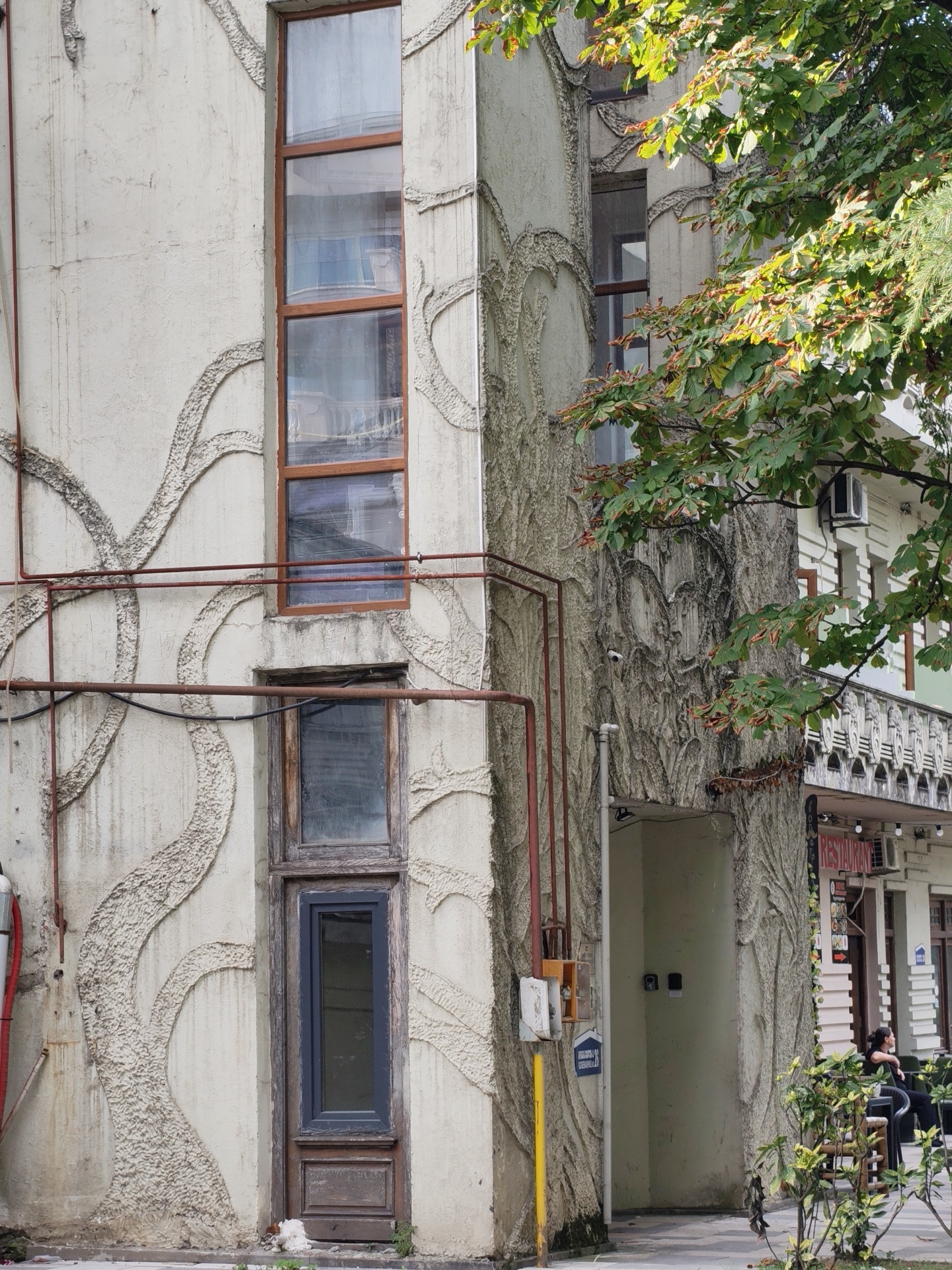The image depicts the exterior of a building with a textured facade resembling tree bark, located in Batumi, Georgia. The building has several tall, narrow windows with wooden frames, some of which appear weathered. Red pipes run vertically and horizontally along the wall. To the right, lush green foliage from a tree drapes over the building. In the background, there is a glimpse of another building with a balcony and signs, including one that reads "RESTAURANT." A person is seated on a bench in front of the restaurant. The overall scene suggests an urban environment with architectural and natural elements.