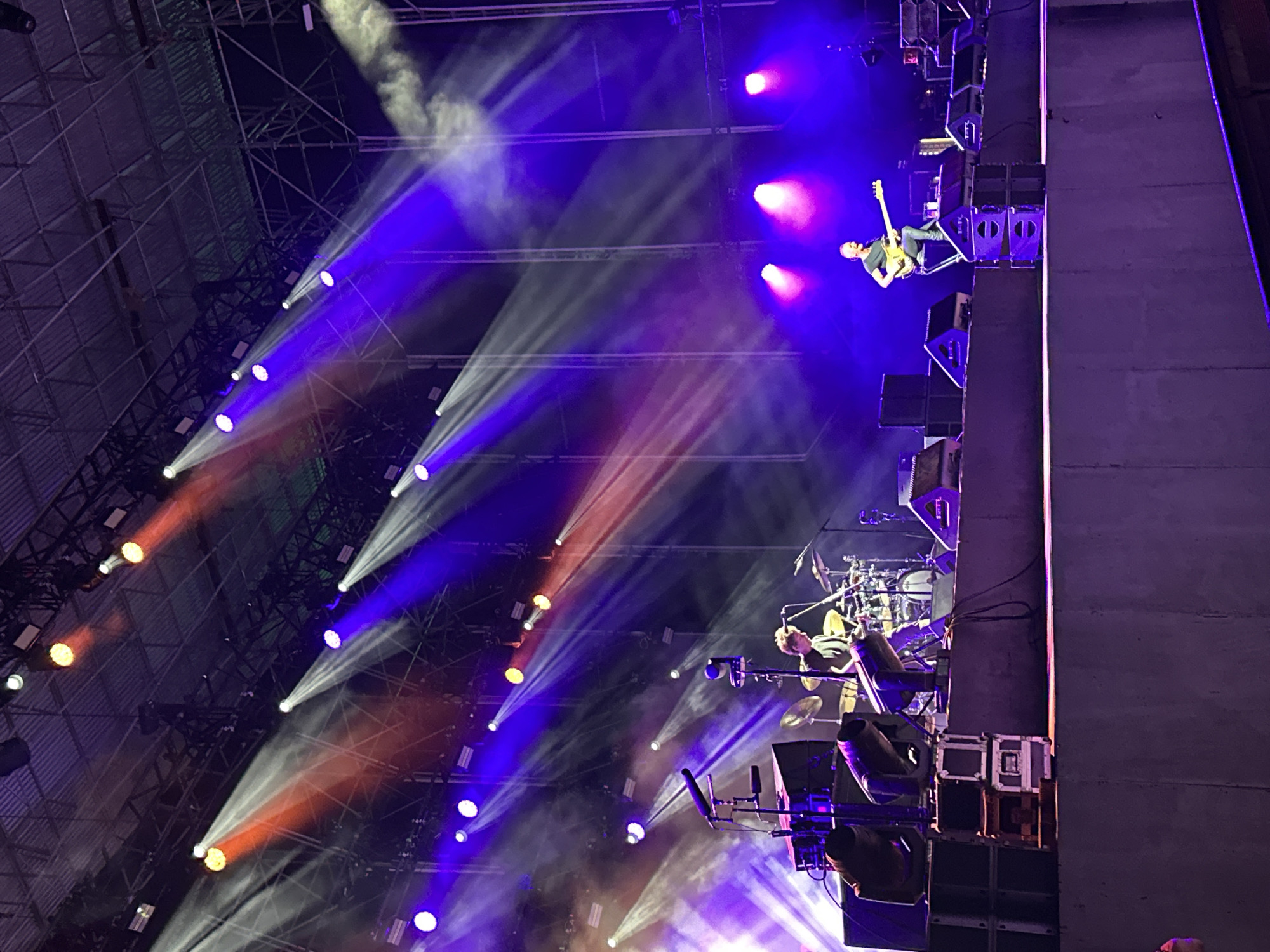 The image depicts an outdoor concert at night in Puerto De Sancti Petri, Spain. A stage is set up with numerous lights casting beams of red and purple light, some of which are partially obscured by smoke. On the stage, two musicians are visible. In the foreground, to the left, a drummer is seated behind a drum kit, playing. To the right, a guitarist is standing, also playing. Both musicians appear focused on their performance. In front of the stage, on a lower level, there are stacks of speakers and lighting equipment. The overall atmosphere suggests a lively musical event.