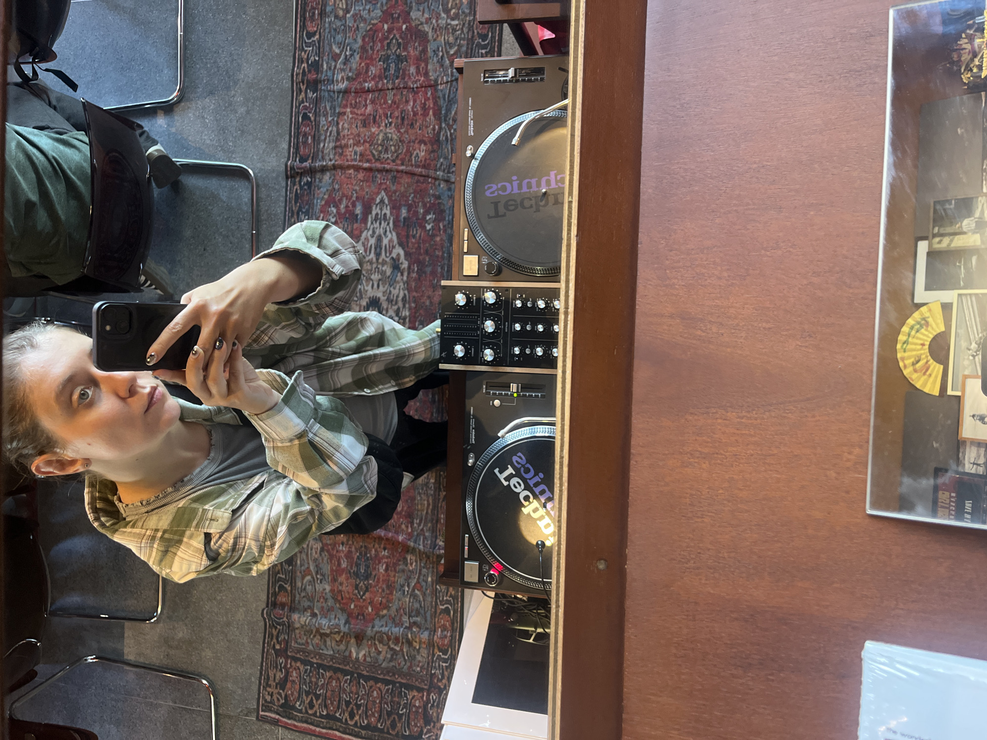 A woman is taking a mirror selfie with a smartphone. She is wearing a green and white plaid flannel shirt and has light skin and shoulder-length hair pulled back. The reflection shows a turntable setup consisting of two Technics turntables and a mixer, mounted in a wooden shelf or stand. To the right of the turntable, there is a framed display of images and a wooden surface. Beneath the turntable, there is visible white packaging. The flooring is gray, and there is a patterned rug visible. Behind the woman, a chair is reflected in the mirror. The location in the city of North Kuta, Indonesia cannot be determined from this image.
