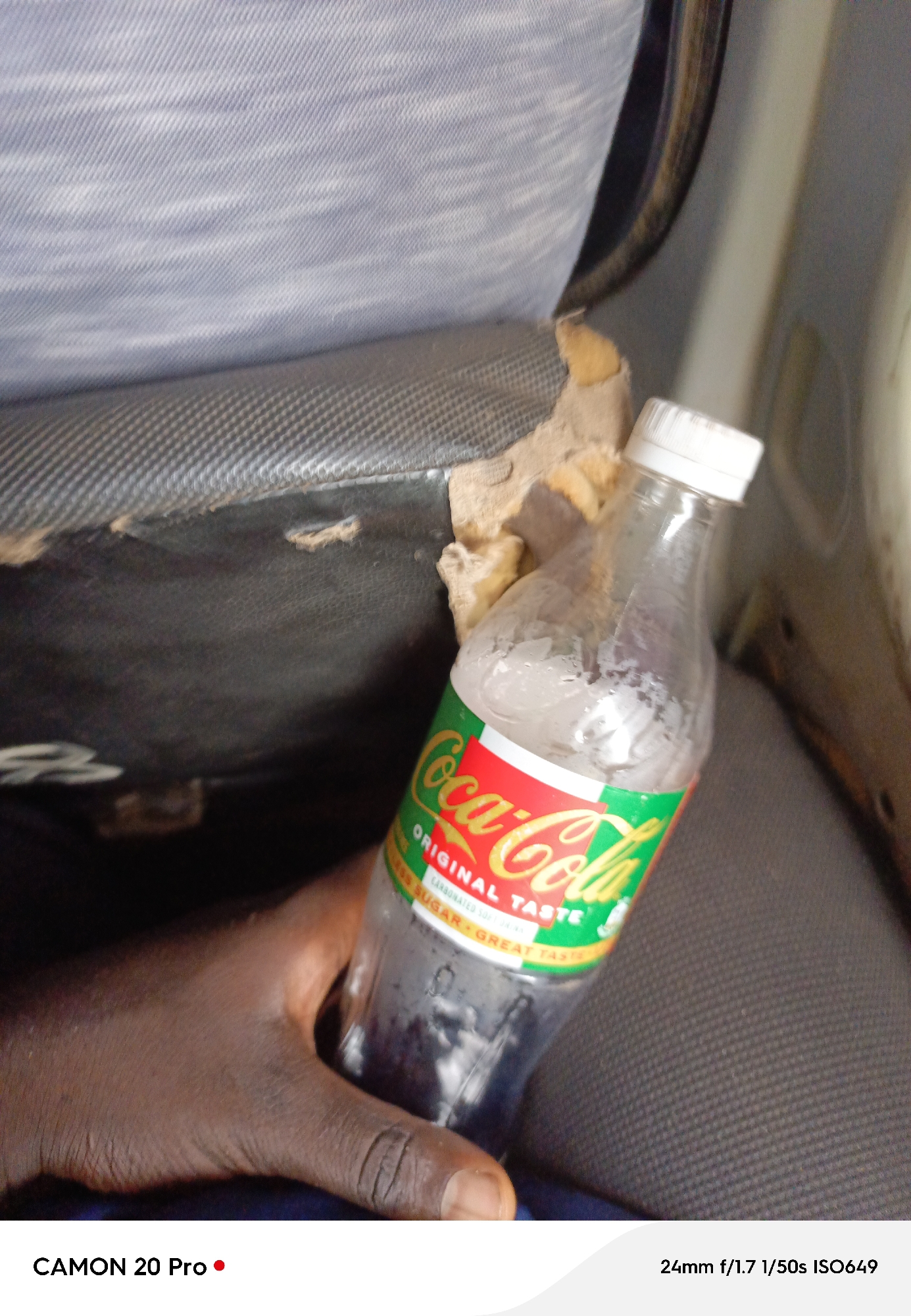 The image shows a close-up of a hand holding a bottle of Coca-Cola. The bottle is partially filled and has condensation on its exterior. The label on the bottle indicates "ORIGINAL TASTE" and "SUGAR · GREAT TASTE". The background appears to be a car interior, with worn upholstery visible. The image is taken with a CAMON 20 Pro, with camera settings of 24mm f/1.7, 1/50s, and ISO649.