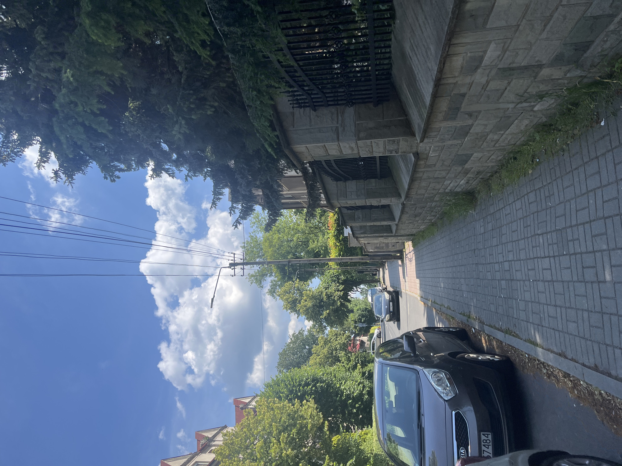 The image depicts a sidewalk and street scene. A grey Kia car is parked on the left side of the street, with its front facing forward. Another car, dark in color, is driving on the right side of the street. The sidewalk is paved with grey bricks. On the right side of the sidewalk, a stone wall with a black metal fence runs along the property line. Tall evergreen trees and deciduous trees line the street and are visible behind the fence. The sky is blue with scattered white clouds. A lamppost is visible on the right side of the street. The buildings on the left side of the street appear to be multi-story residential buildings with red roofs. The overall environment suggests a suburban or residential area within a city.