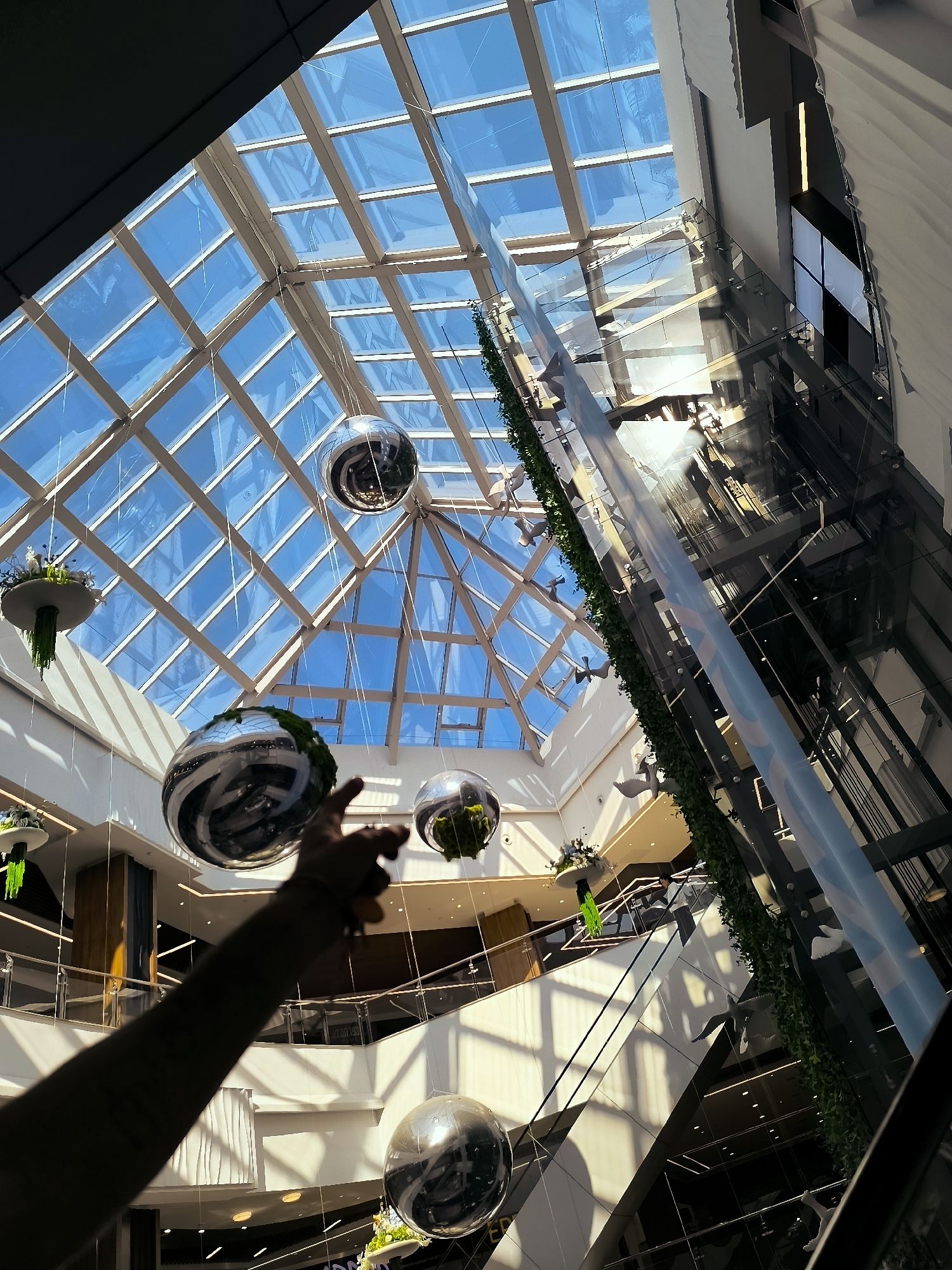 The image depicts the interior of a modern, multi-level shopping mall with a large, glass-paneled ceiling. Sunlight streams through the geometric pattern of the ceiling, casting strong shadows and highlights throughout the atrium. Several large, reflective spherical ornaments, some adorned with greenery, are suspended from the ceiling by thin wires, creating a sense of scale and visual interest. A glass elevator shaft, encased by a vertical living wall of green foliage, rises prominently through the space. On the escalators and walkways of the upper levels, several individuals can be seen, some interacting and others appearing to be moving through the mall. In the foreground, a person's arm and hand are visible, with the index finger extended as if pointing at one of the hanging spheres. The architecture suggests a contemporary commercial environment designed to maximize natural light.