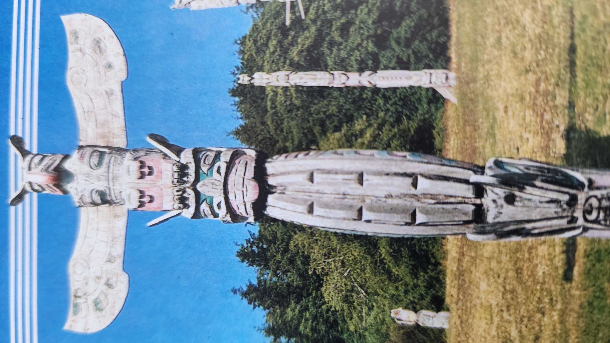 This is a medium shot of a totem pole in a field with a forest in the background under a clear blue sky. The totem pole in the foreground is tall and carved from wood with intricate designs and figures. It is mostly painted white, with some touches of red, green, and pink. The pole has an owl at the top with outstretched wings, followed by a human-like face with large eyes and a mouth. Below that is another carved face with green and black markings, and the rest of the pole is carved with rectangular shapes and abstract patterns. There are other totem poles in the background, one of which is partially visible and appears to be a similar style but is further away and less detailed. The field is covered in dry grass, and the forest consists of tall, dark green coniferous trees. The image is slightly blurry, giving it a vintage postcard-like appearance. The location is not specified in the image.