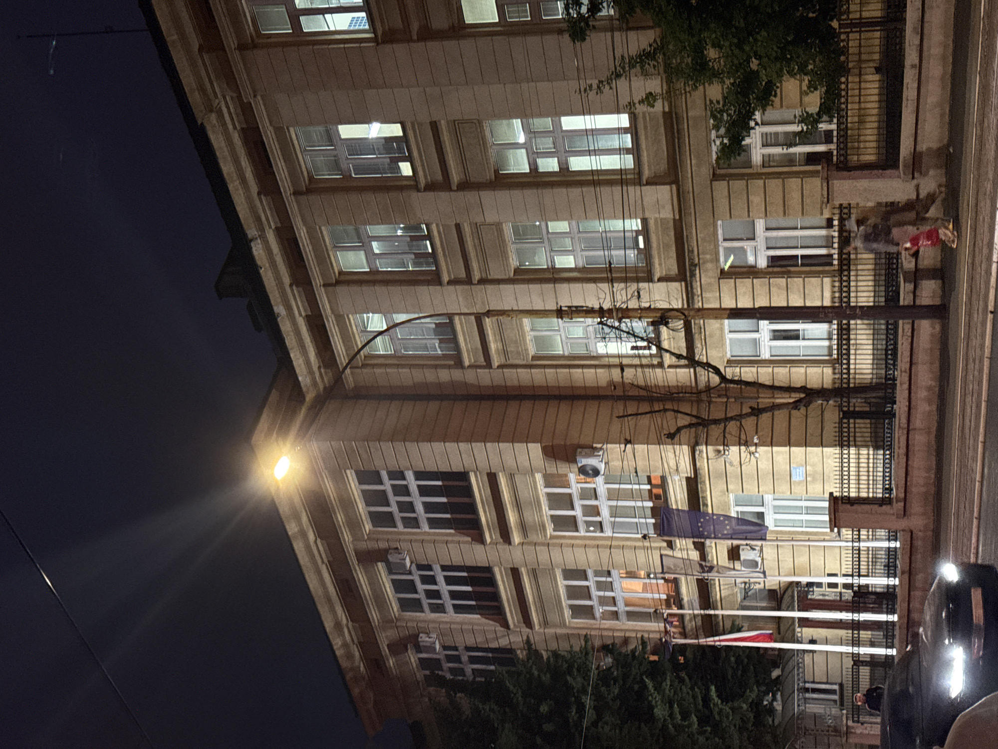 The image depicts a multi-story building at night in Belgrade, Serbia. The building is illuminated by lights inside and a street lamp nearby. There are many windows, most of which have white frames. The building's facade is light brown, and it has a classical architectural style with decorative molding around the windows. Flags are affixed to the building's exterior; one appears to be the European Union flag, and another could be the Serbian flag. A wrought-iron fence runs along the front of the building at street level. A tree is in the foreground, partially obscuring the view. A car is parked along the street, its headlights visible. A person is walking along the sidewalk. Wires stretch across the front of the building.