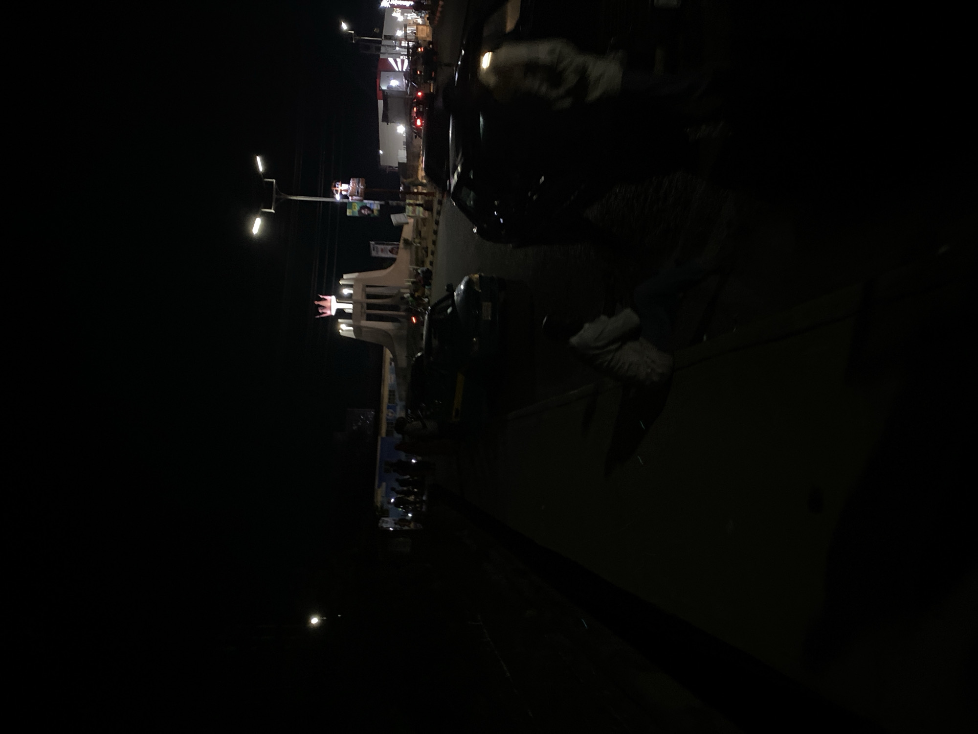 At night in Barakin Lerawi, Nigeria, the image captures a street scene. The architecture features a lit gateway structure, potentially marking a significant location. A group of people are assembled near the gateway. Streetlights illuminate the scene, with cars visible on the road. Additional structures, possibly businesses, are indicated by lights further down the road.