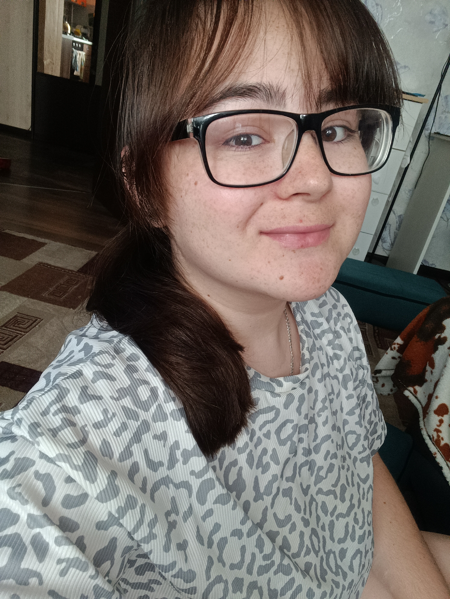 A young woman with brown hair and glasses is pictured from the chest up, taking a selfie. She is indoors, with a carpet visible on the floor and furniture in the background. The woman is wearing a light-colored shirt with a grey leopard print pattern. The background suggests a residential setting, possibly an apartment. There are no discernable elements within the image that indicate a specific location in Noviy, Russia.