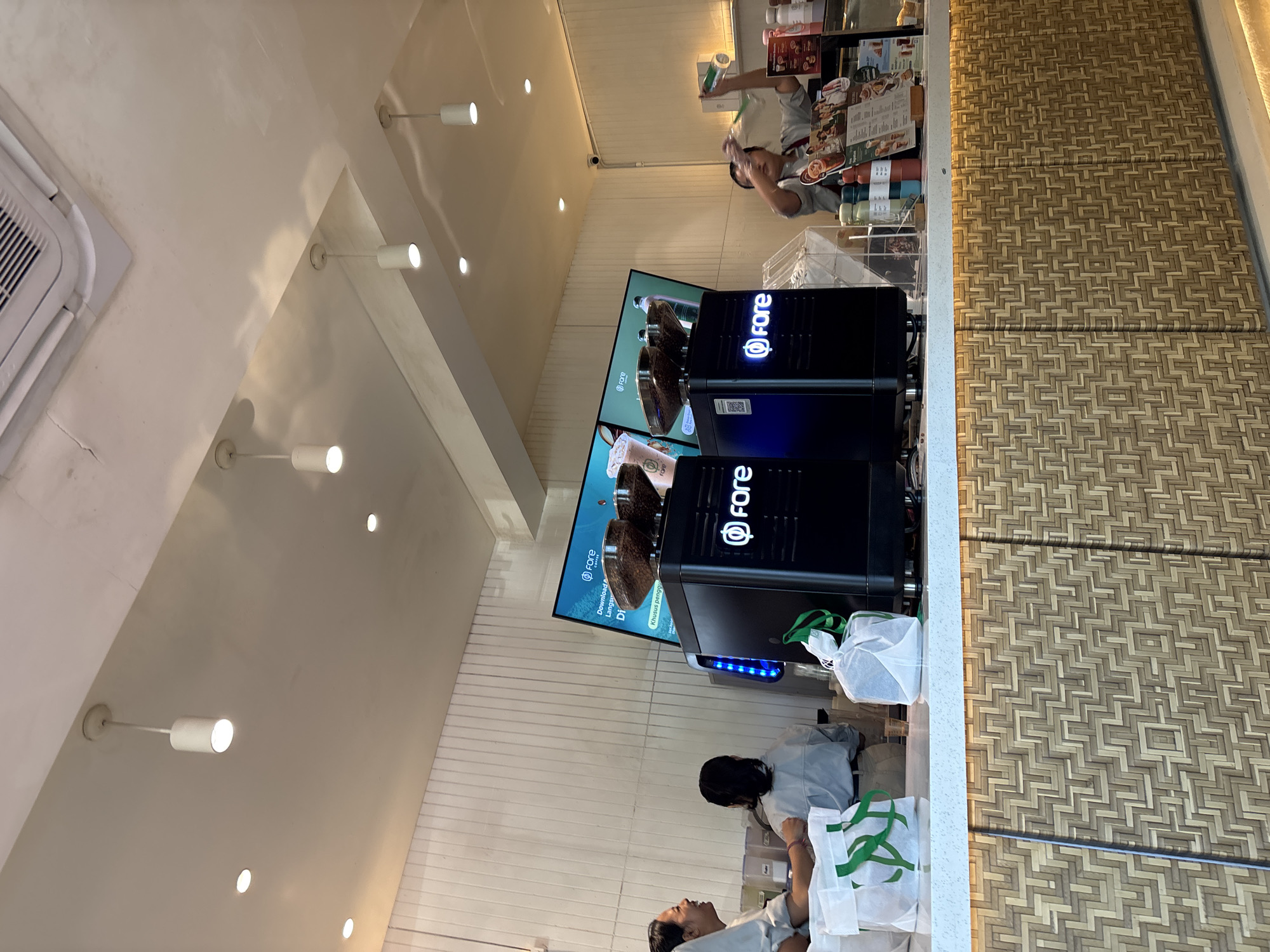 The image captures the interior of a coffee shop, likely Fore Coffee, in Denpasar, Indonesia. Two baristas are visible behind the counter, one in the process of preparing a drink and another interacting with a customer. A prominent display screen shows the Fore Coffee logo and promotional material. Two black coffee machines with the Fore Coffee logo are positioned on the counter, each topped with a hopper filled with coffee beans. Several paper bags with green Fore Coffee branding are also on the counter. The counter itself is decorated with a woven bamboo pattern, and overhead, recessed lighting illuminates the space, along with several pendant lights. The overall atmosphere suggests a busy, modern coffee shop environment.
