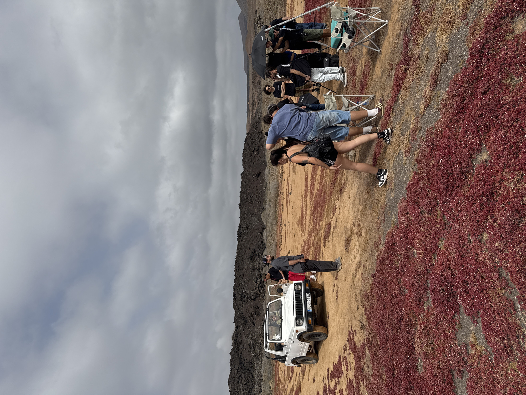 The image depicts a film crew and tourists in a barren, rocky landscape, likely a volcanic area in Tinajo, Spain. The ground is covered in patches of red ground cover. A white Suzuki SUV is parked near a rocky, dark-colored land formation. Two men lean against the vehicle. Nearby, a couple walks across the landscape. Further away, the film crew is assembled, some sitting in chairs under an umbrella. The sky is overcast with gray clouds.