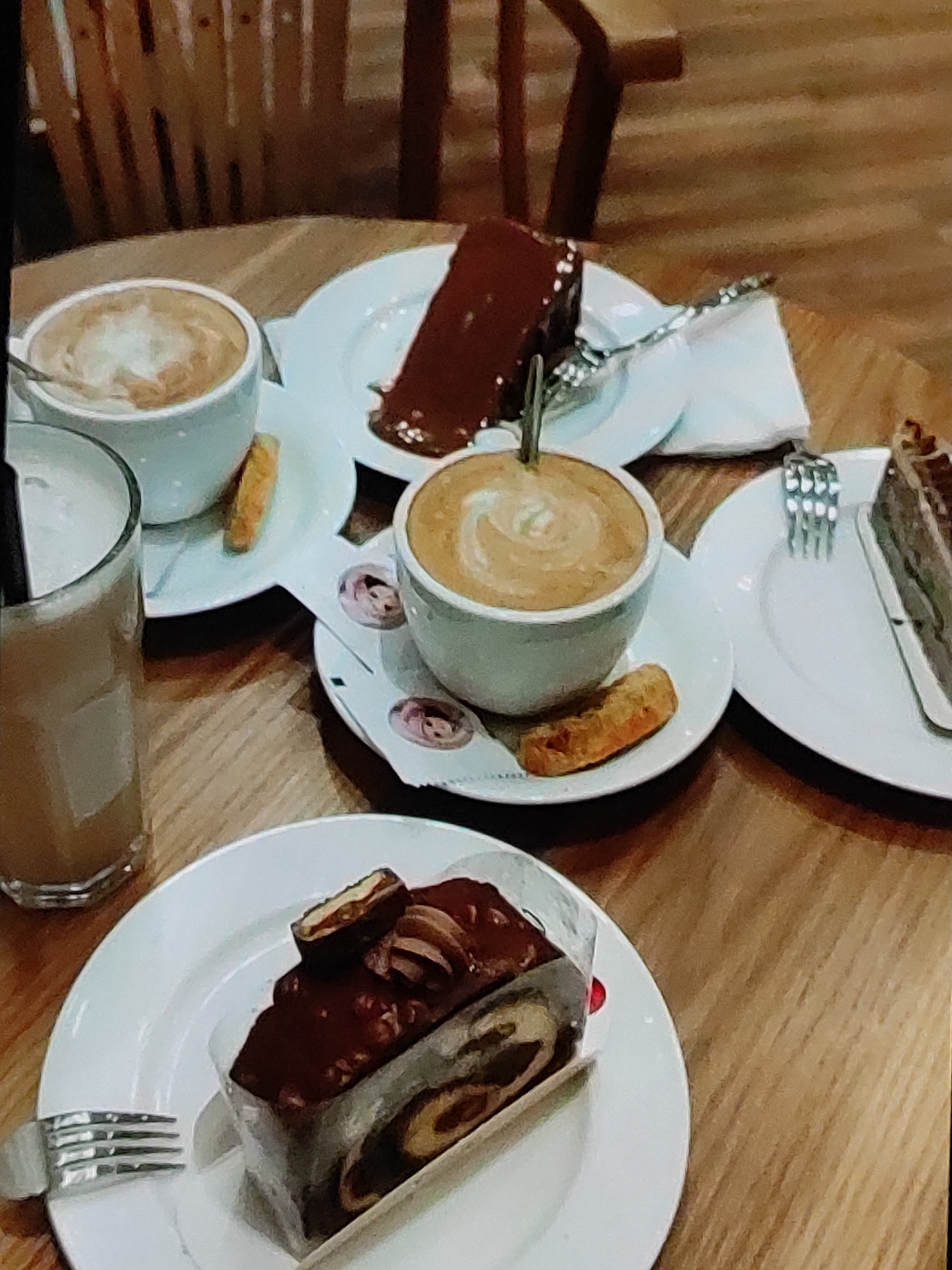This is a medium shot, eye-level, slightly out-of-focus, interior photograph of a table setting in a cafe. The table is made of light brown wood with a visible grain. On the table are several plates of desserts and cups of coffee. In the foreground, a slice of chocolate cake with a swirl pattern inside sits on a white plate, topped with two pieces of what appears to be a chocolate bar. A fork rests to the left of the plate. To the left of the cake is a tall glass of a light brown beverage with a black straw. Behind this, on another white plate, is a cup of coffee with latte art. A small, orange, rectangular biscuit is on the saucer. Further back and to the right, a rectangular slice of dark chocolate cake is on a white plate. Another white cup of coffee with latte art is situated to the left of this cake, with a small orange biscuit on its saucer. A silver fork is placed to the right of the chocolate cake. In the background, wooden chair backs are visible. The overall lighting is warm and diffused. The scene appears to depict a cafe in Tashkent, Uzbekistan, likely a dessert or coffee shop, given the popular offerings.