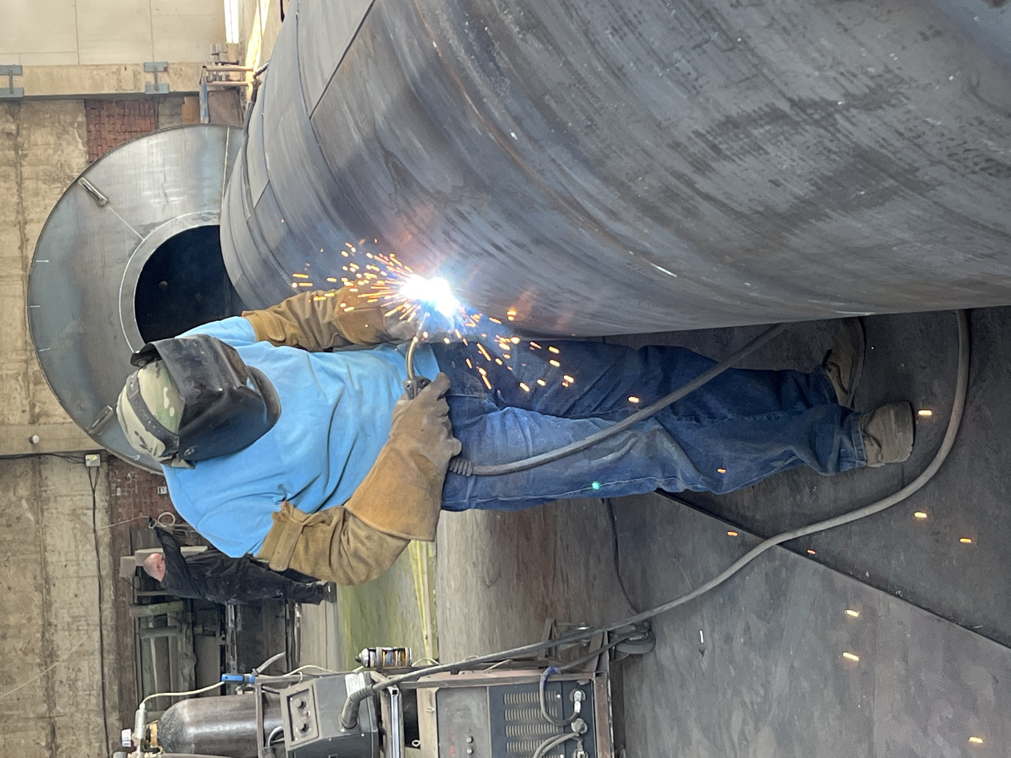 A welder, wearing a protective mask, helmet, blue shirt, tan gloves, and blue jeans, is actively welding a large cylindrical object. Sparks are flying from the welding torch. The scene takes place in an industrial setting, possibly a workshop or factory, with concrete flooring and large metal structures visible in the background. A welding machine and a gas cylinder are present to the left of the welder. In the background, another person in dark clothing is visible, facing away from the camera and appearing to be engaged in work. The environment suggests a manufacturing or repair facility, consistent with a location in Taganrog City District, Russia.
