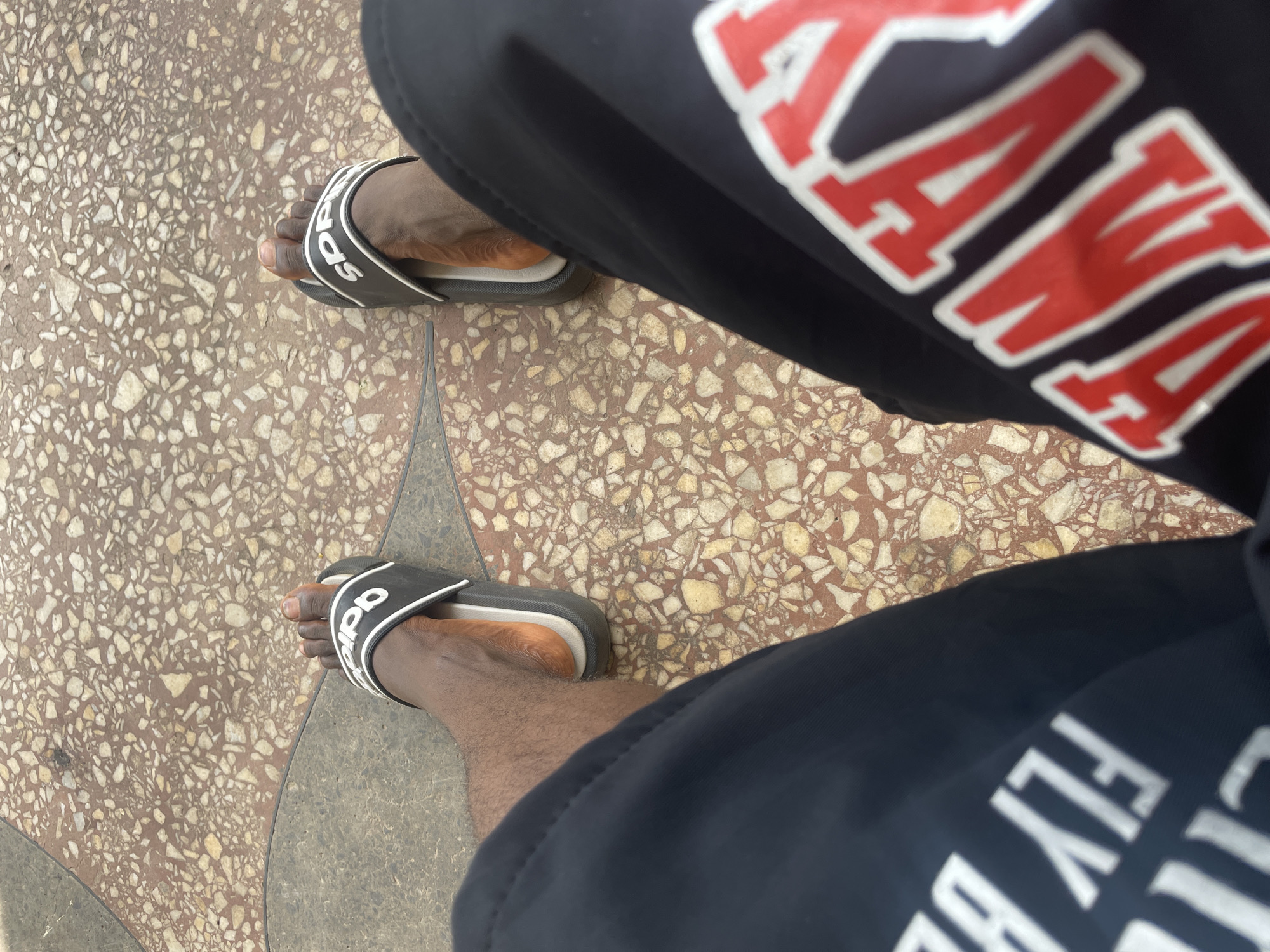 This is a first-person, high-angle shot of a person's legs and feet, wearing dark blue shorts with white and red lettering and black Adidas slide sandals. The person is sitting on a textured ground composed of small, irregular stones in shades of brown, tan, and white, with darker gray grout lines. The scene is set outdoors, likely in Jos, Nigeria, given the warm, dry appearance of the ground and the ambient light. The ground appears to be a paved walkway or patio. The writing on the shorts is partially visible; on the left leg, white lettering reads "FLY BR", and on the right leg, red lettering with a white outline reads "ANT".