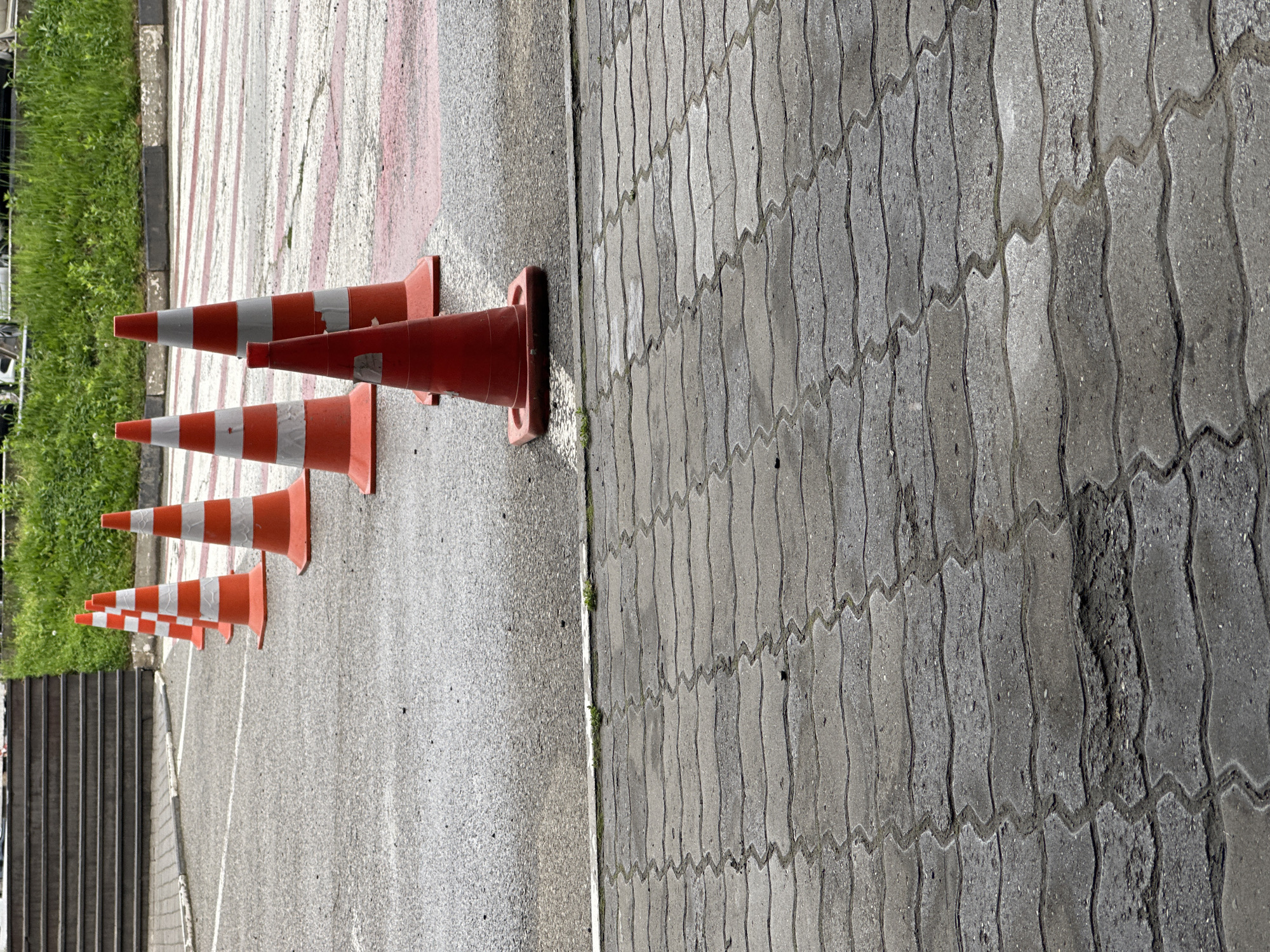 The image depicts an outdoor urban scene. A row of orange and white striped traffic cones are lined up on a concrete surface, next to a section of gray, wavy-patterned paving stones. The paving stones show signs of wear and discoloration. To the left of the cones, there is a set of concrete stairs and a small patch of greenery. A red and white striped crosswalk-like area is visible in the background behind the cones. The city of Unknown, Unknown is not discernible from the image.
